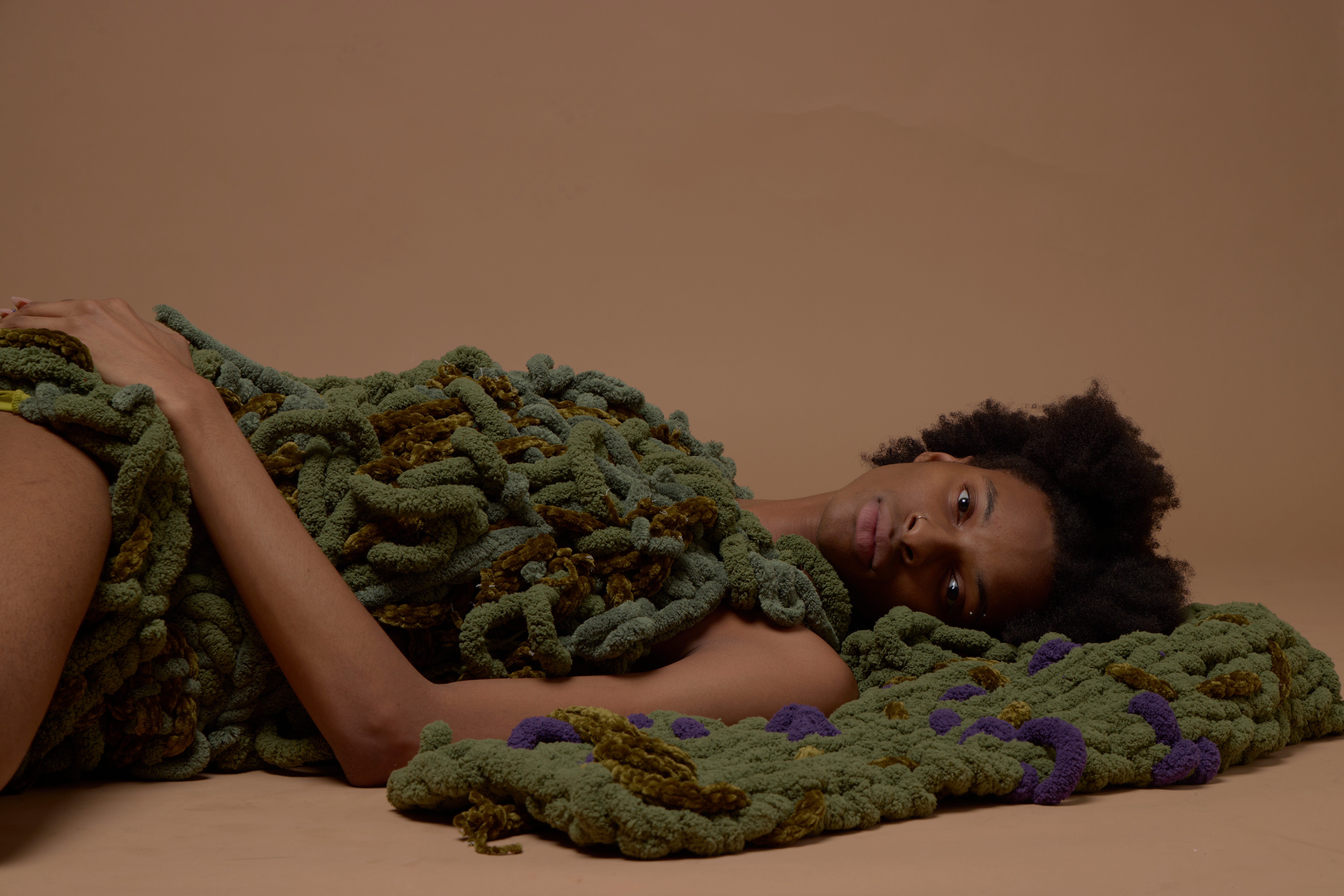 Person lying on a surface covered with textured, colorful fabric, looking at the camera with a neutral expression.