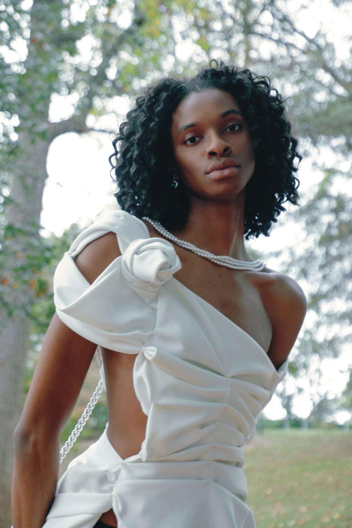 Woman with curly hair in a white dress outdoors, trees in background.