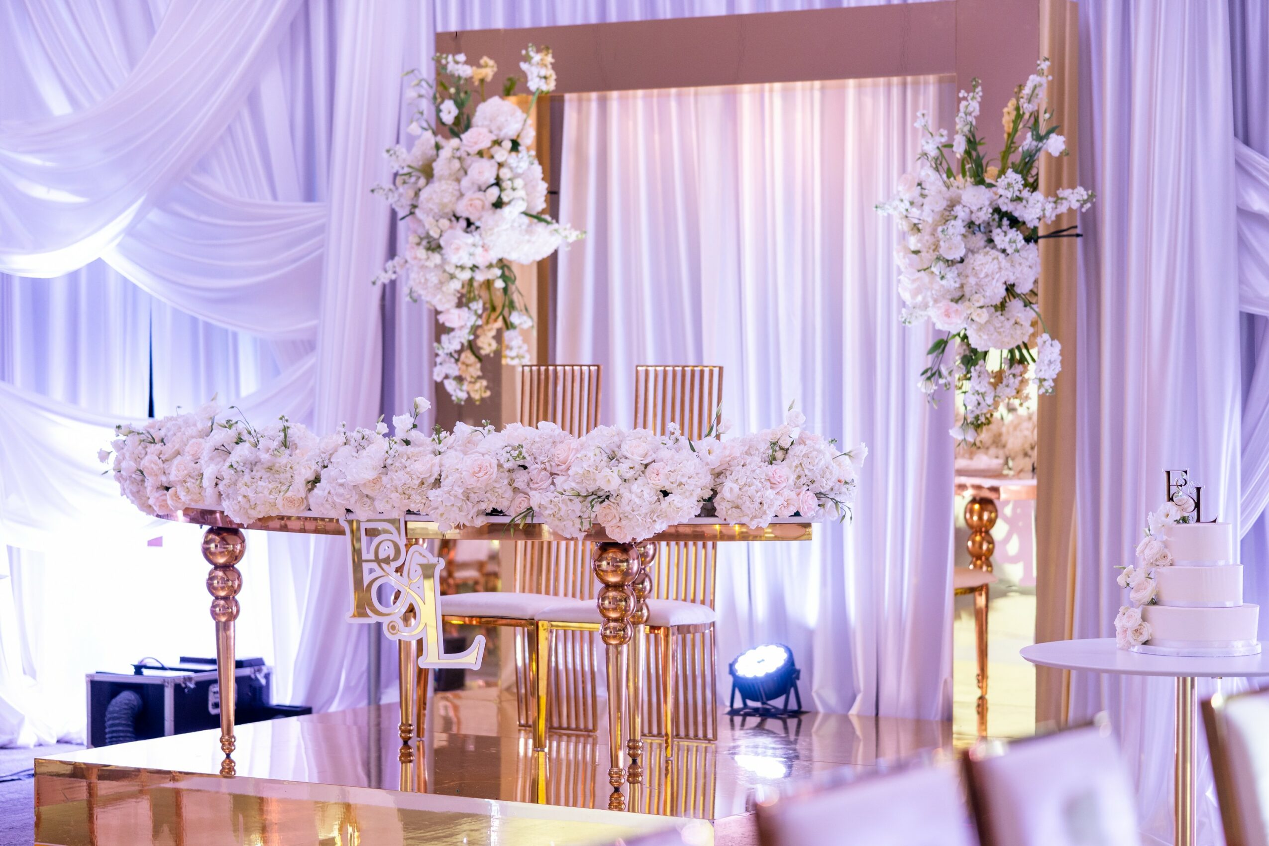 Elegant wedding or event setup with floral arrangements, draped curtains, and a decorated table with chairs, in soft pastel colors.
