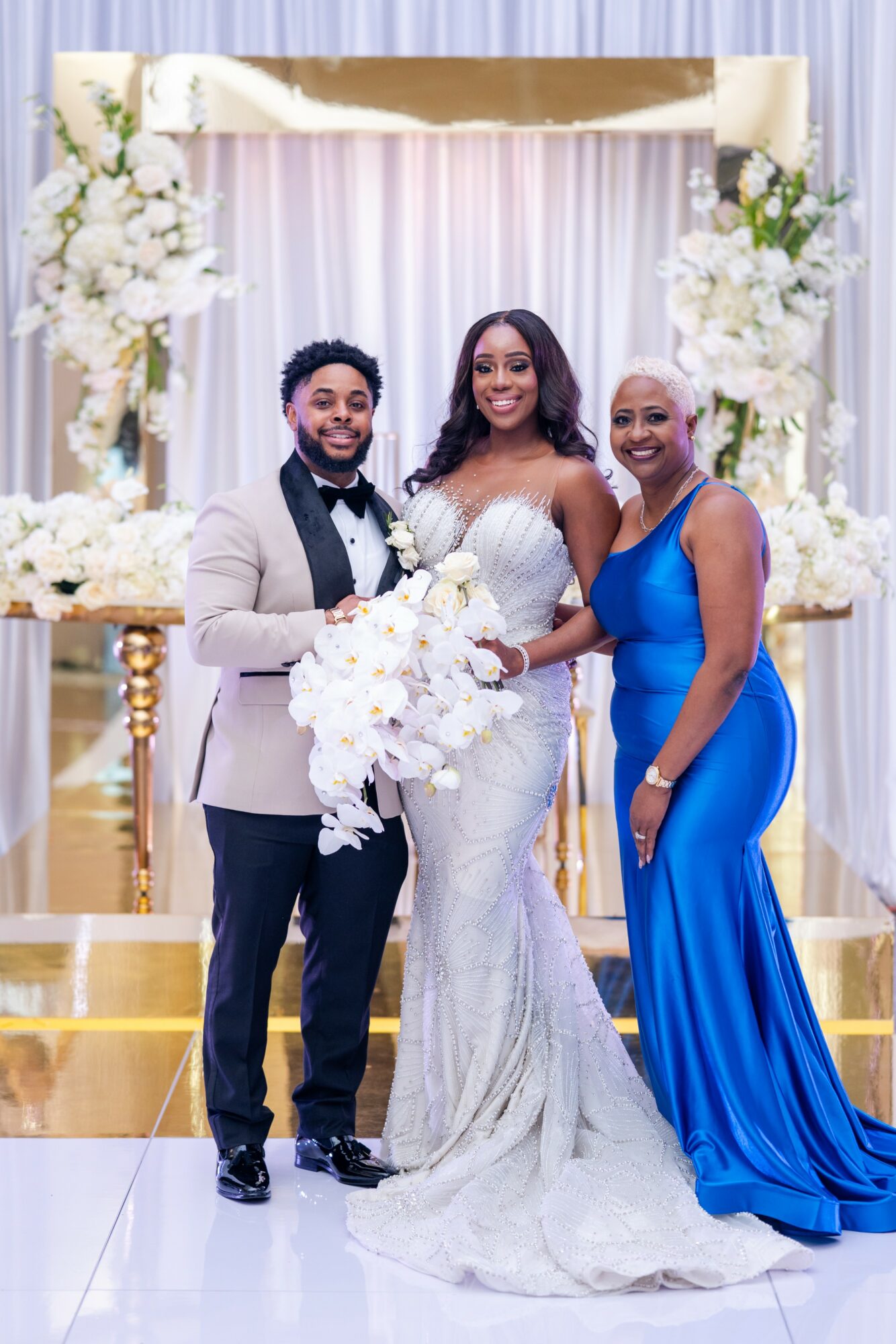 Three people in formal attire standing in front of a decorated backdrop with flowers, smiling at the camera.