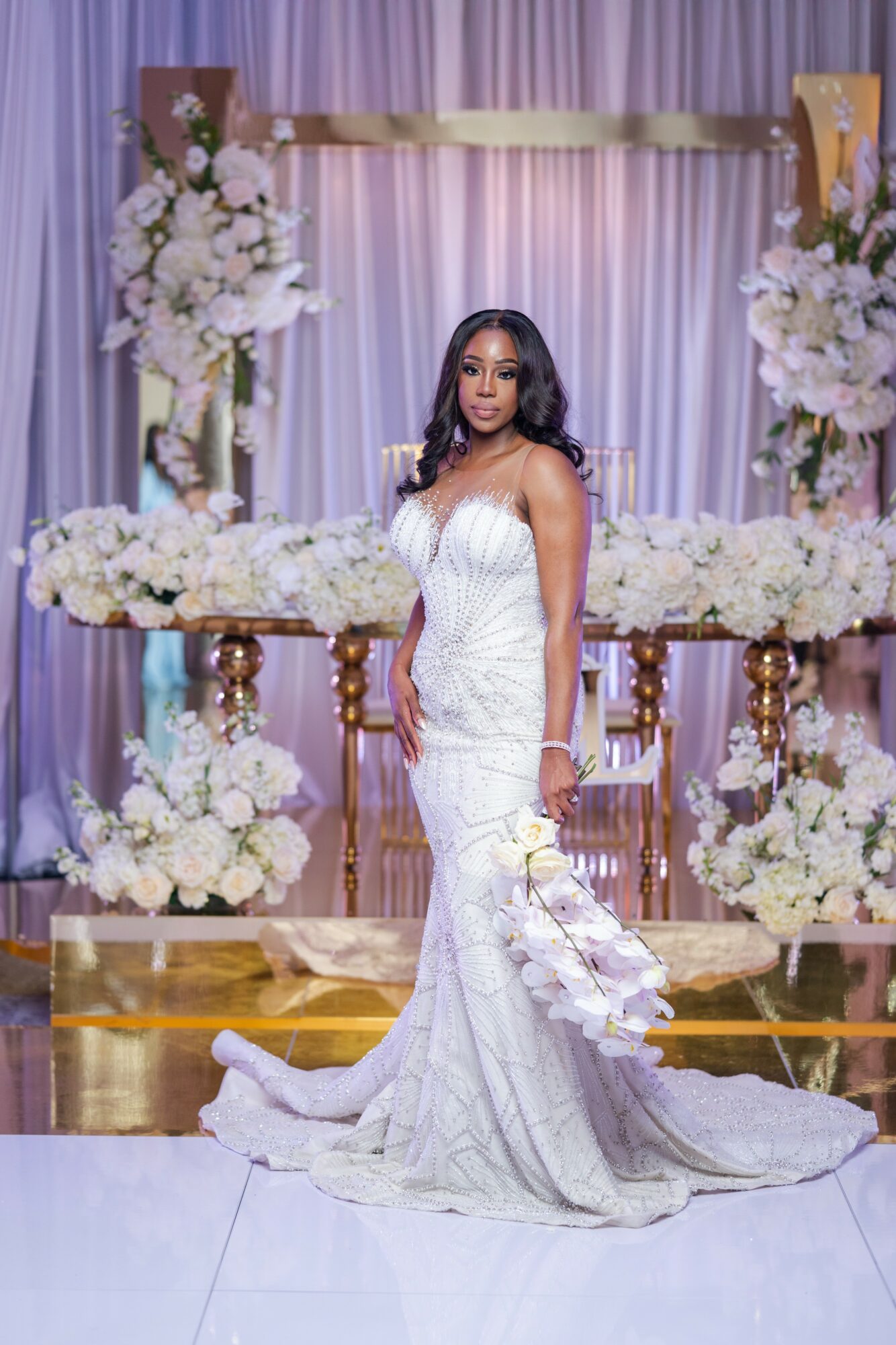 Woman in a white wedding dress standing on a decorated stage with floral arrangements and drapery.