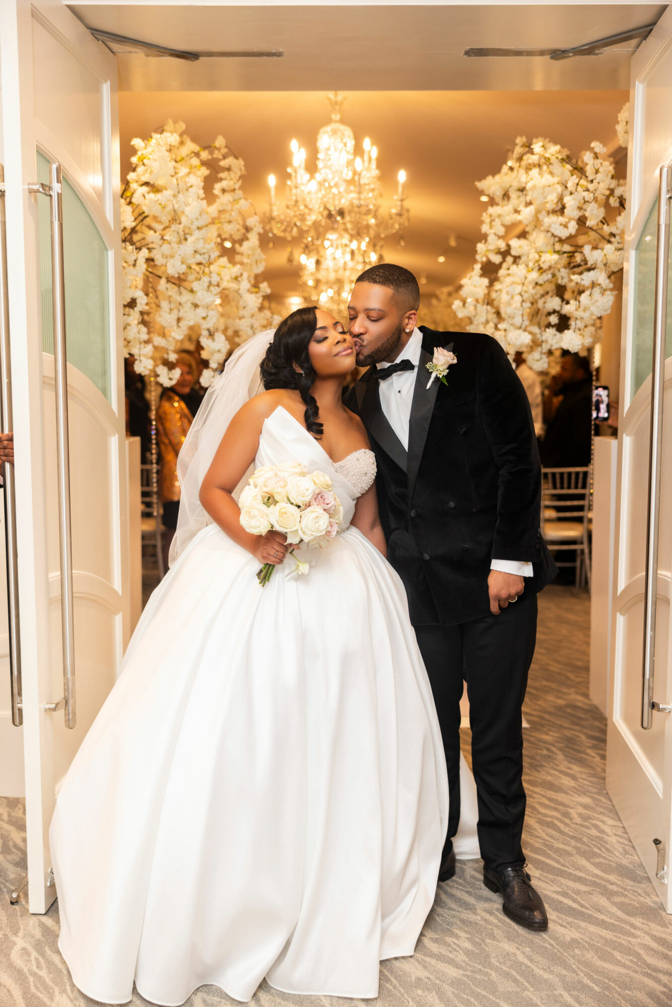 Bride and groom kissing in a decorated wedding venue with chandeliers and floral arrangements, bride holding a bouquet, groom in tuxedo.