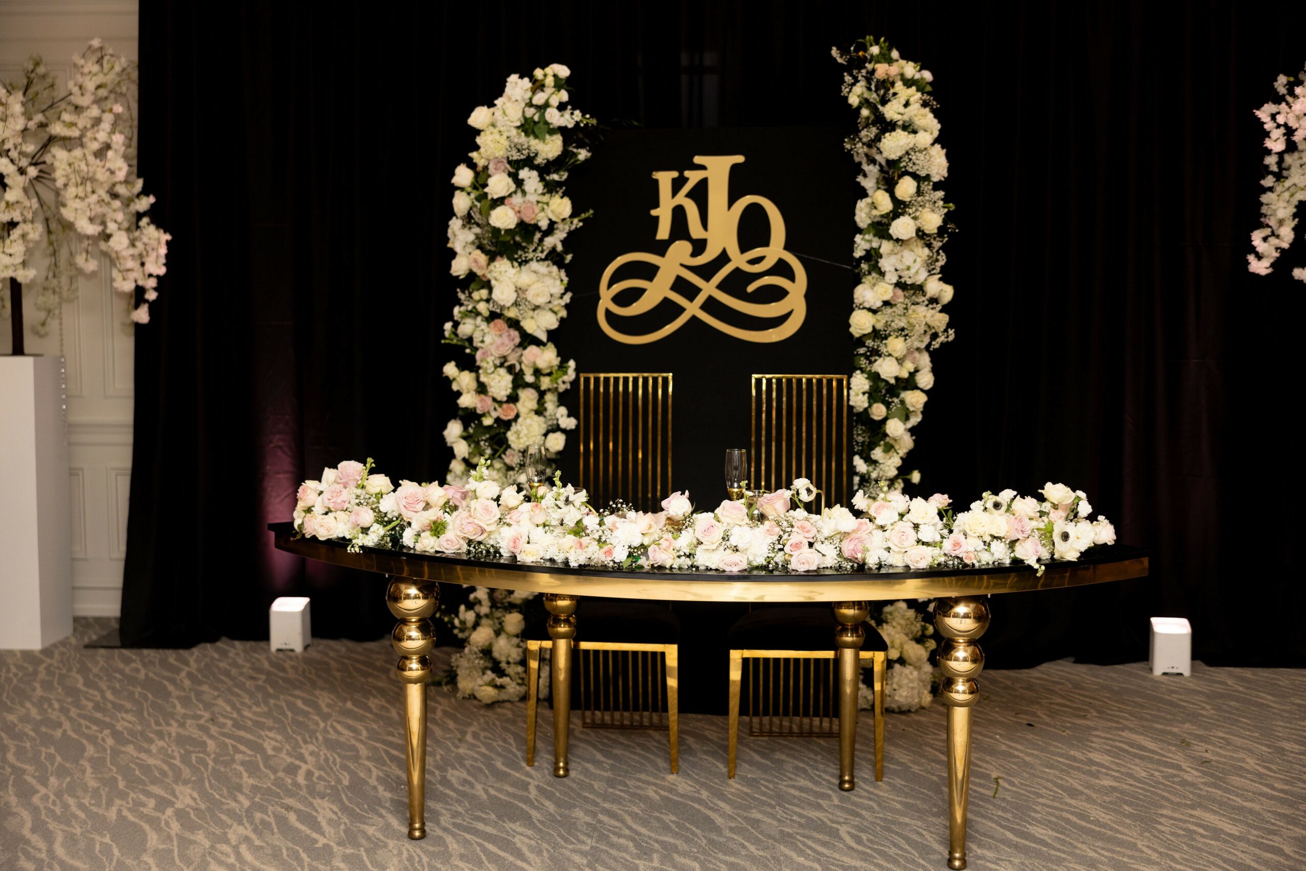 Elegant wedding or event table with floral arrangements and a monogram backdrop, gold chairs, and dark curtains.