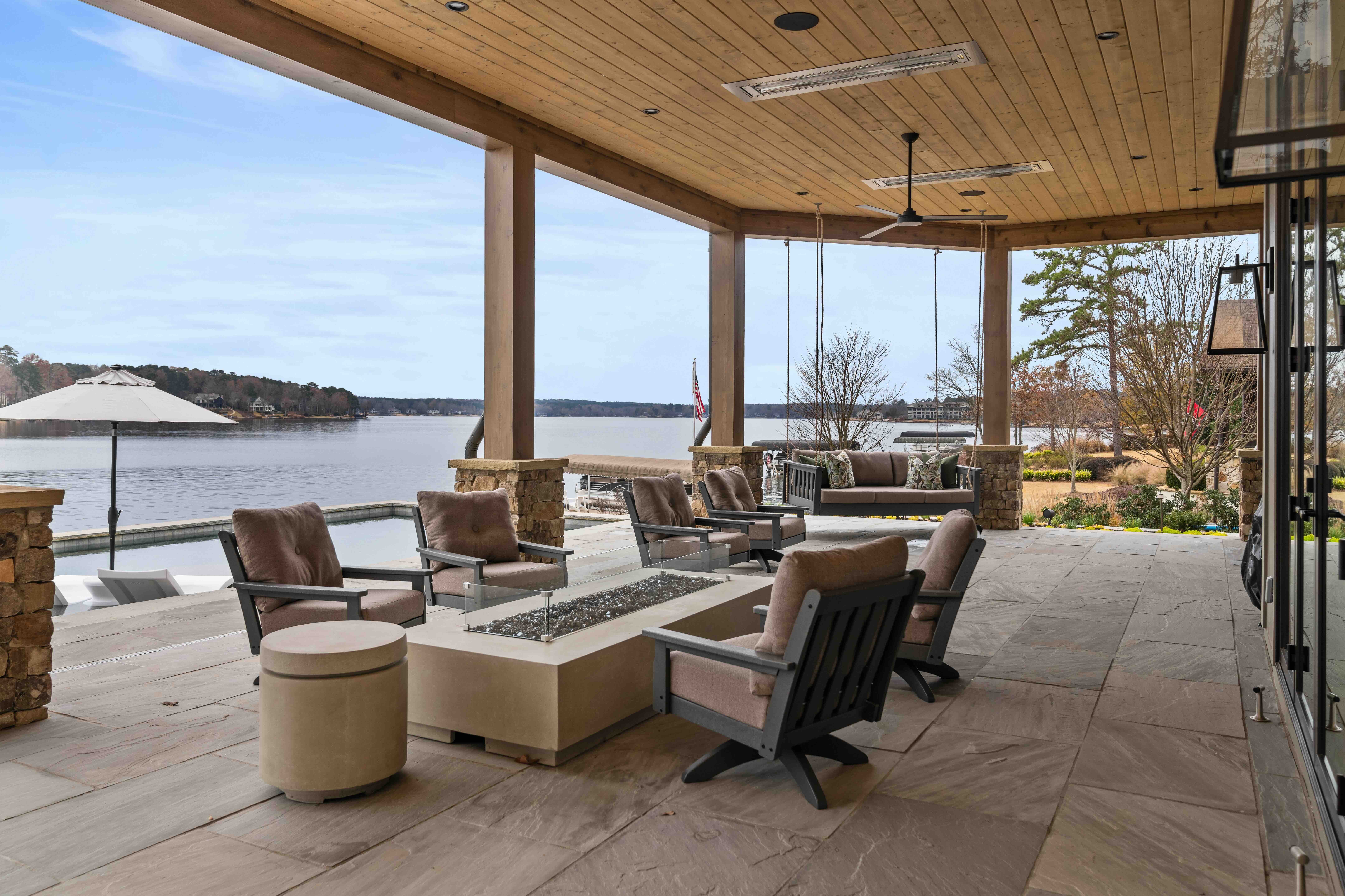 Indoor seating area with chairs and tables overlooking a lake through large glass windows, with outdoor scenery and trees visible.
