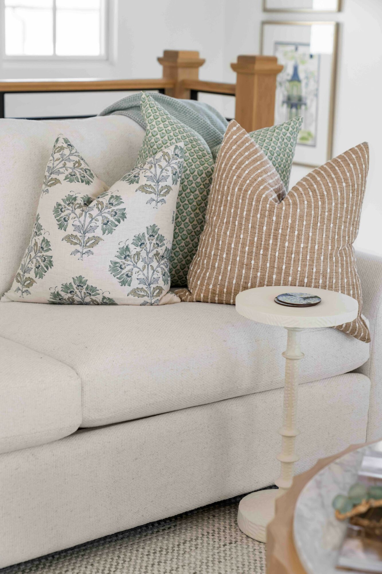 Four decorative pillows on a light-colored sofa with a small round side table nearby, in a bright room.