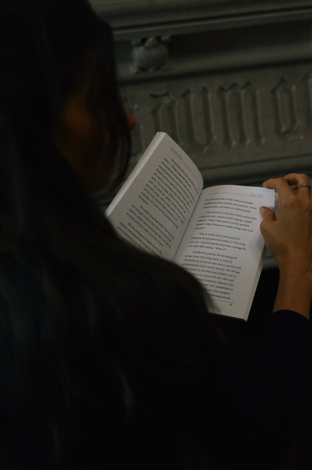 Person reading a book with text visible, sitting near a radiator, with dark hair and a dark top.
