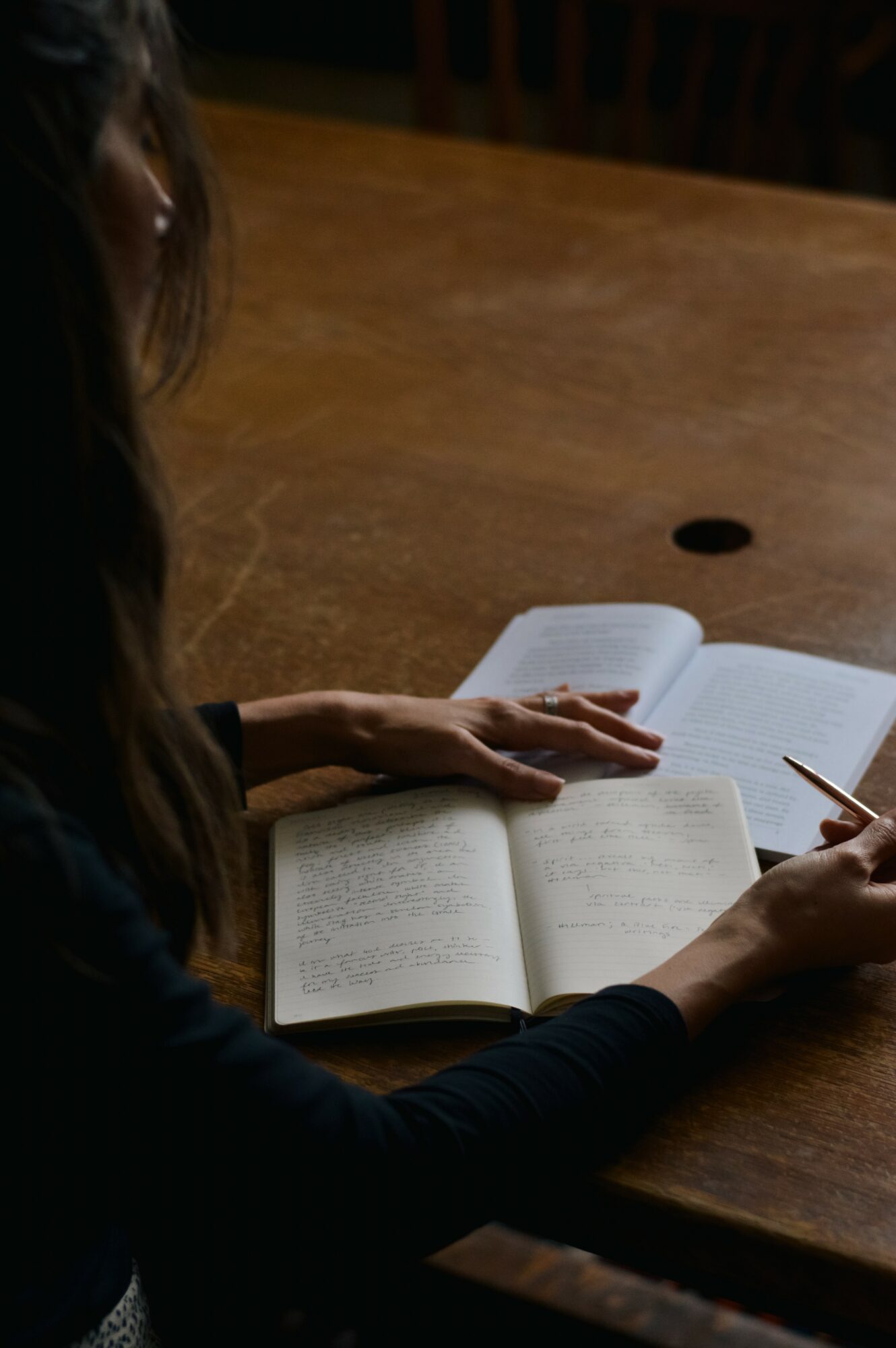 Person reading a book at a wooden table, with a pen in hand, in a dimly lit room.