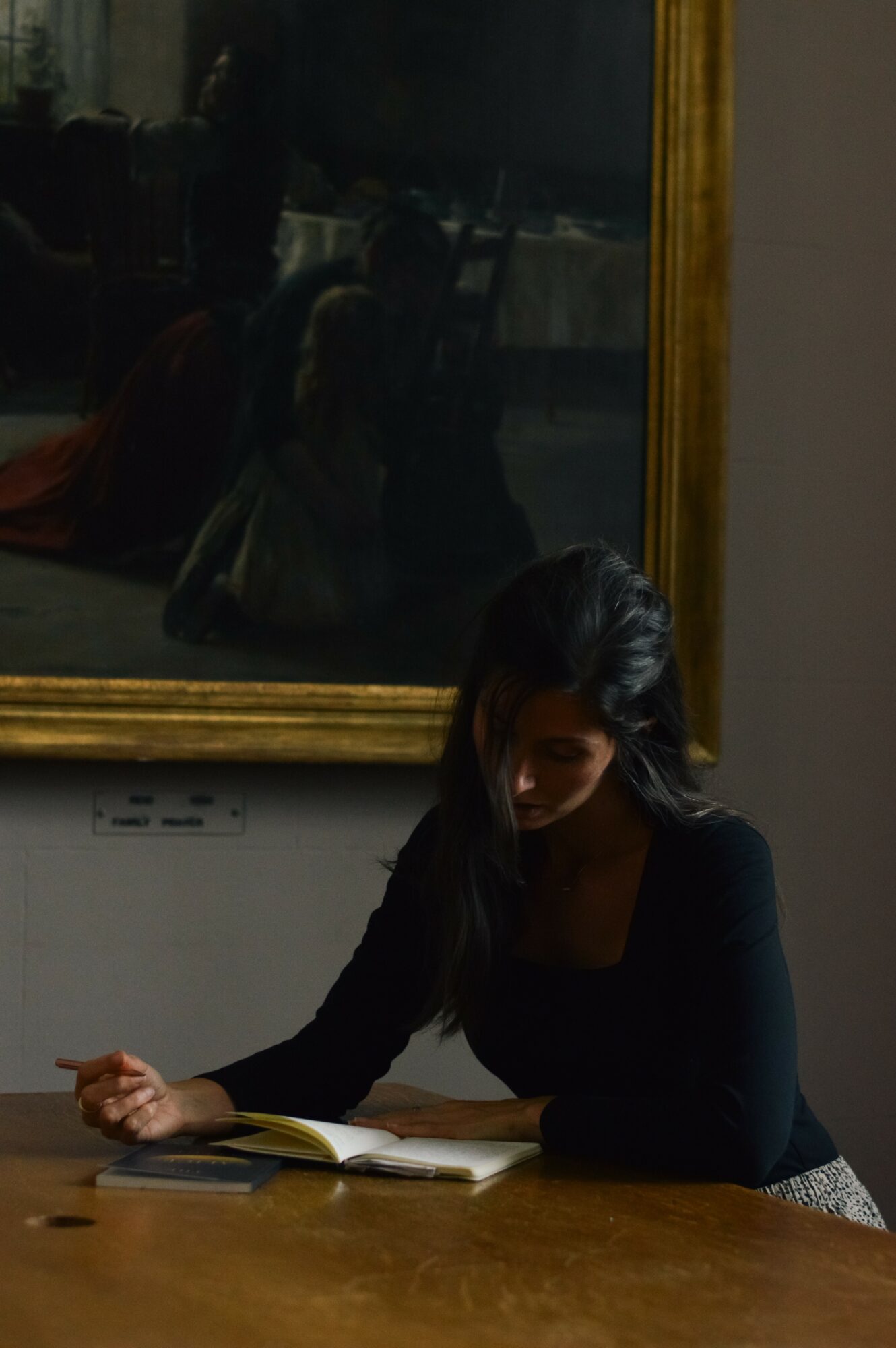 Woman with dark hair reading a book at a table, reflection visible in a large mirror behind her.