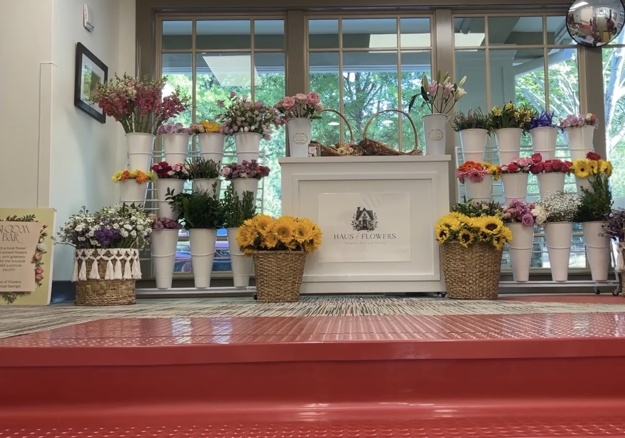 Display of colorful flowers in vases and baskets on a windowsill and floor, with a white sign in the center.