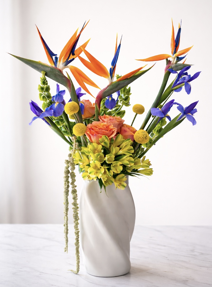 Colorful flower arrangement with orange, purple, yellow, and green flowers in a white vase on a white surface.