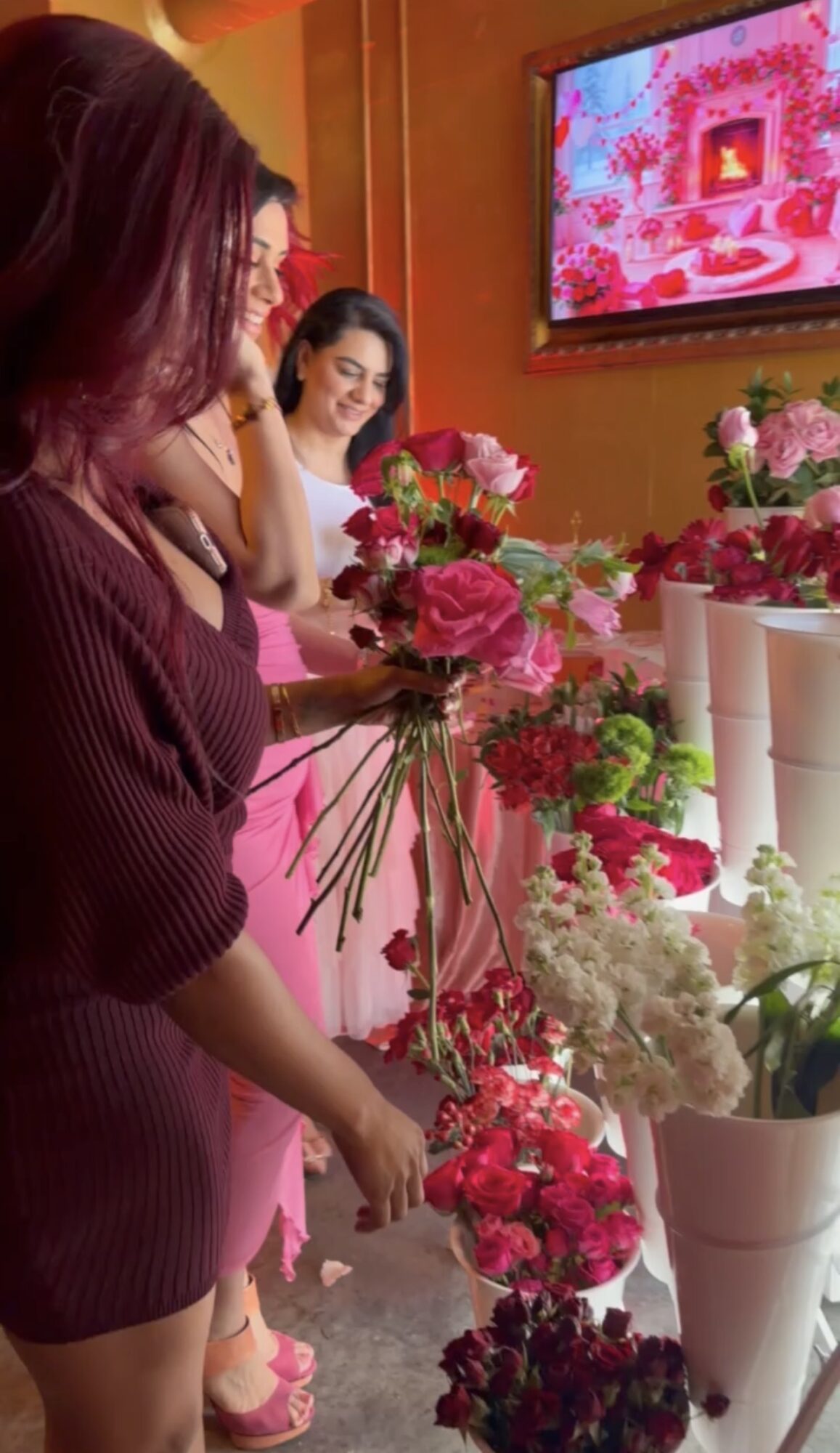 Two women with flowers in a shop, one smiling, pink and red flowers on display, TV screen in background.