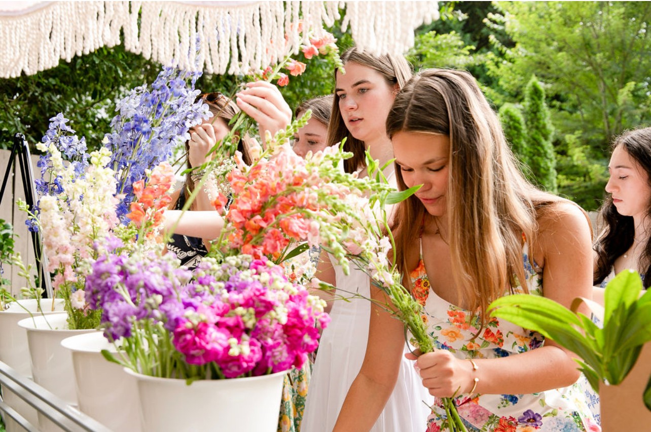 Group of women looking at colorful flowers outdoors, surrounded by greenery and potted plants.