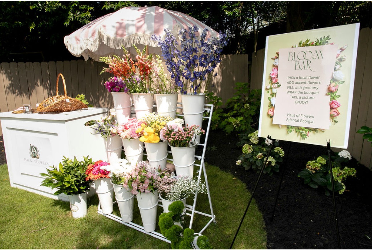 Flower display with white buckets, umbrella, and a sign in a garden setting.