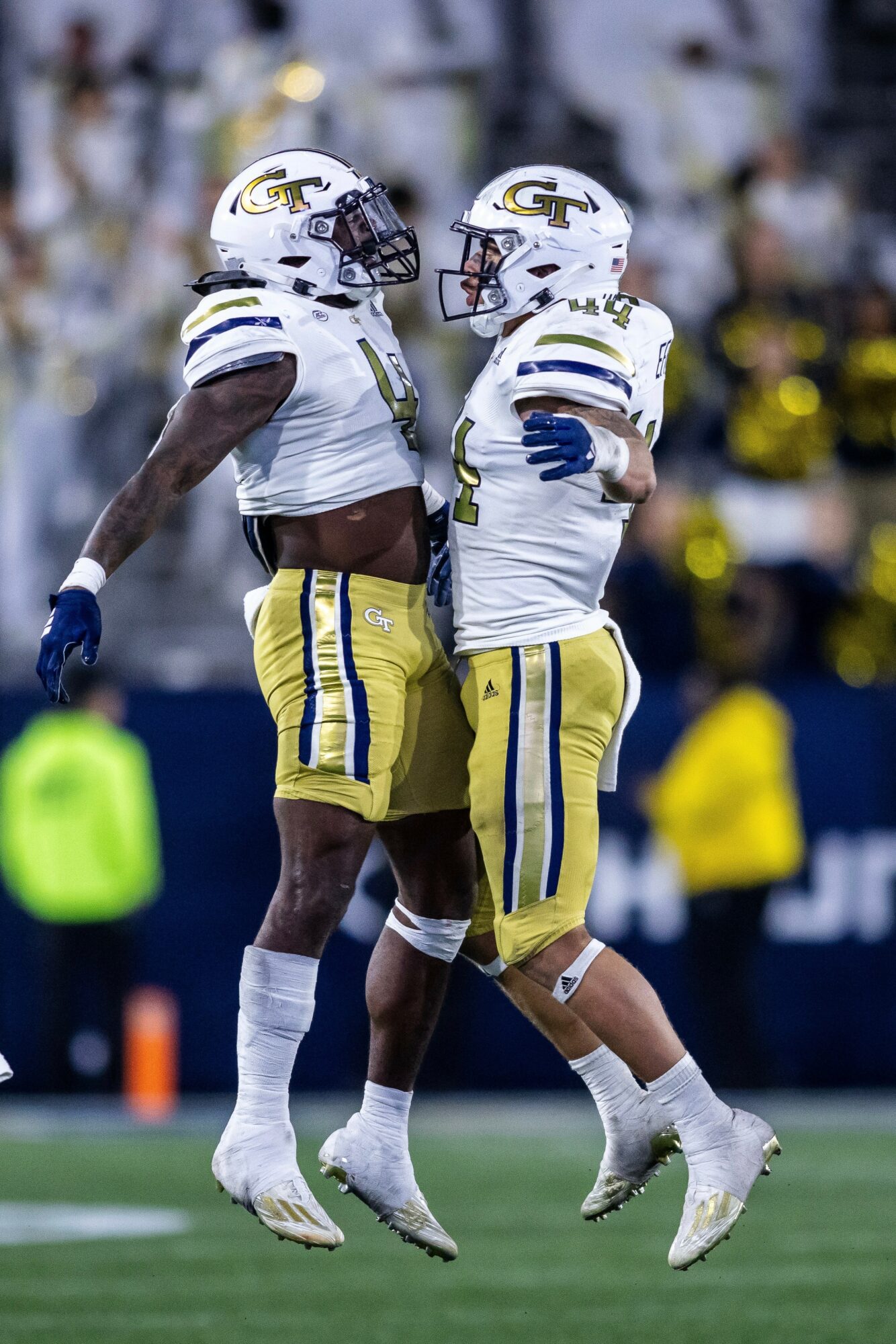Two football players in white and yellow uniforms celebrate on the field, jumping and facing each other.
