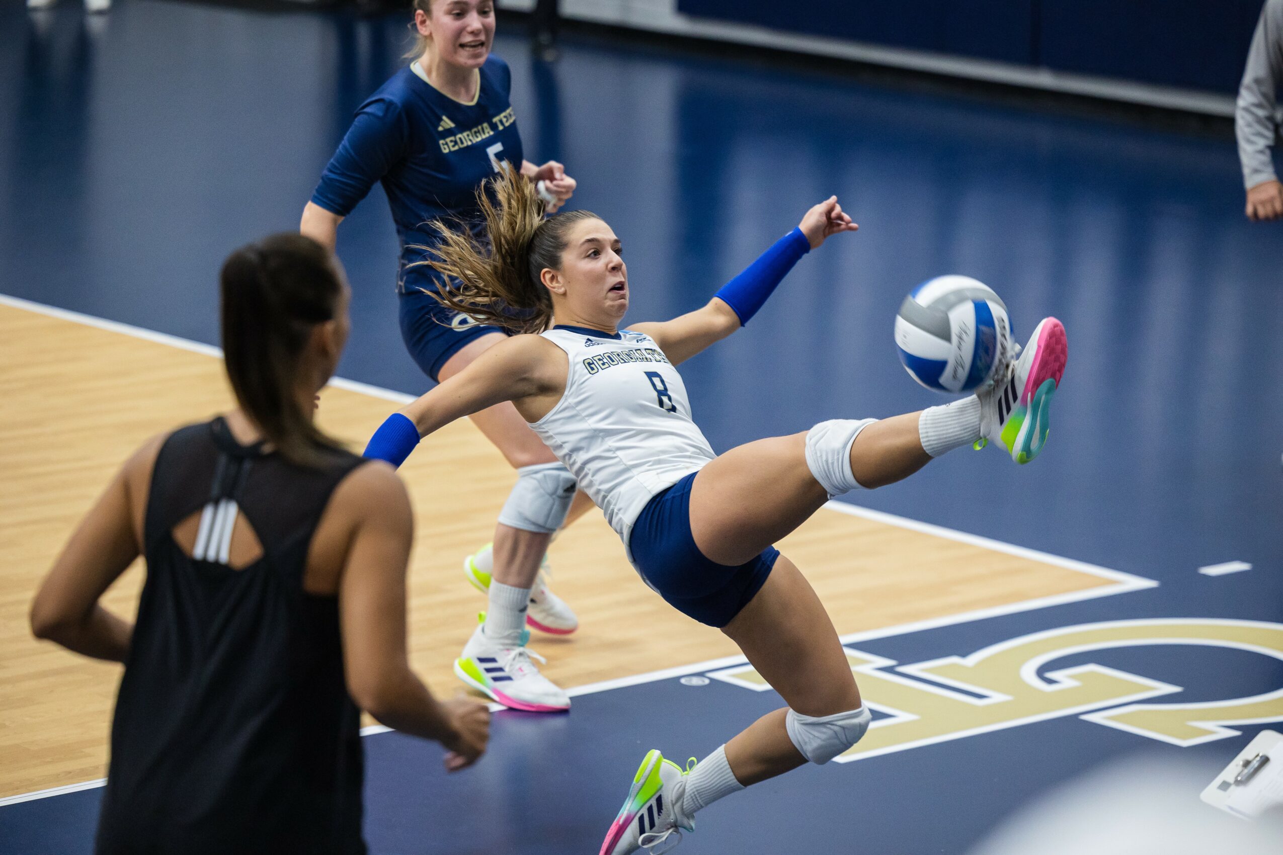 A female volleyball player in mid-air attempting a spike during a game, with other players nearby on the court.