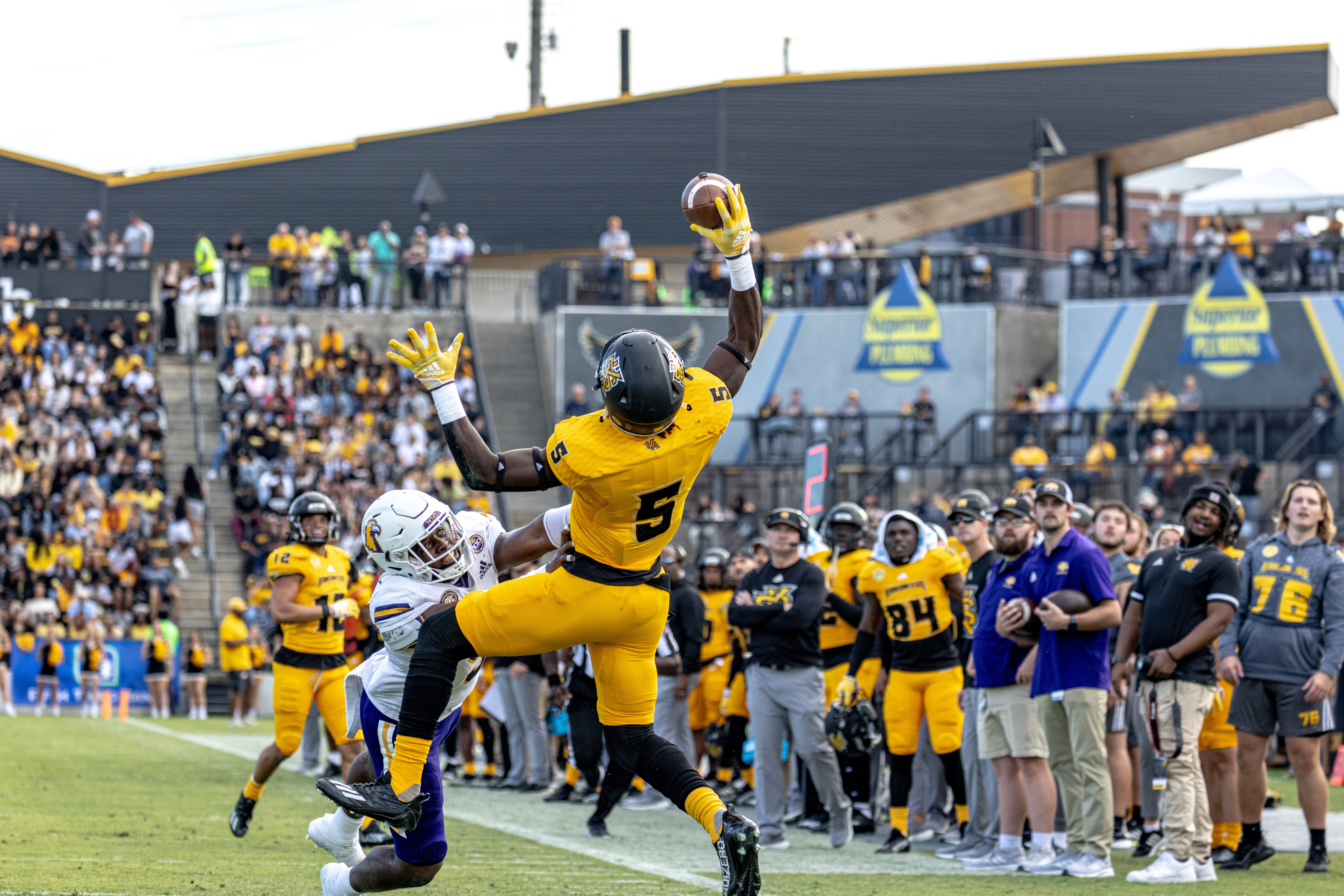 Football player in yellow uniform catches the ball in a stadium with spectators and team members nearby.