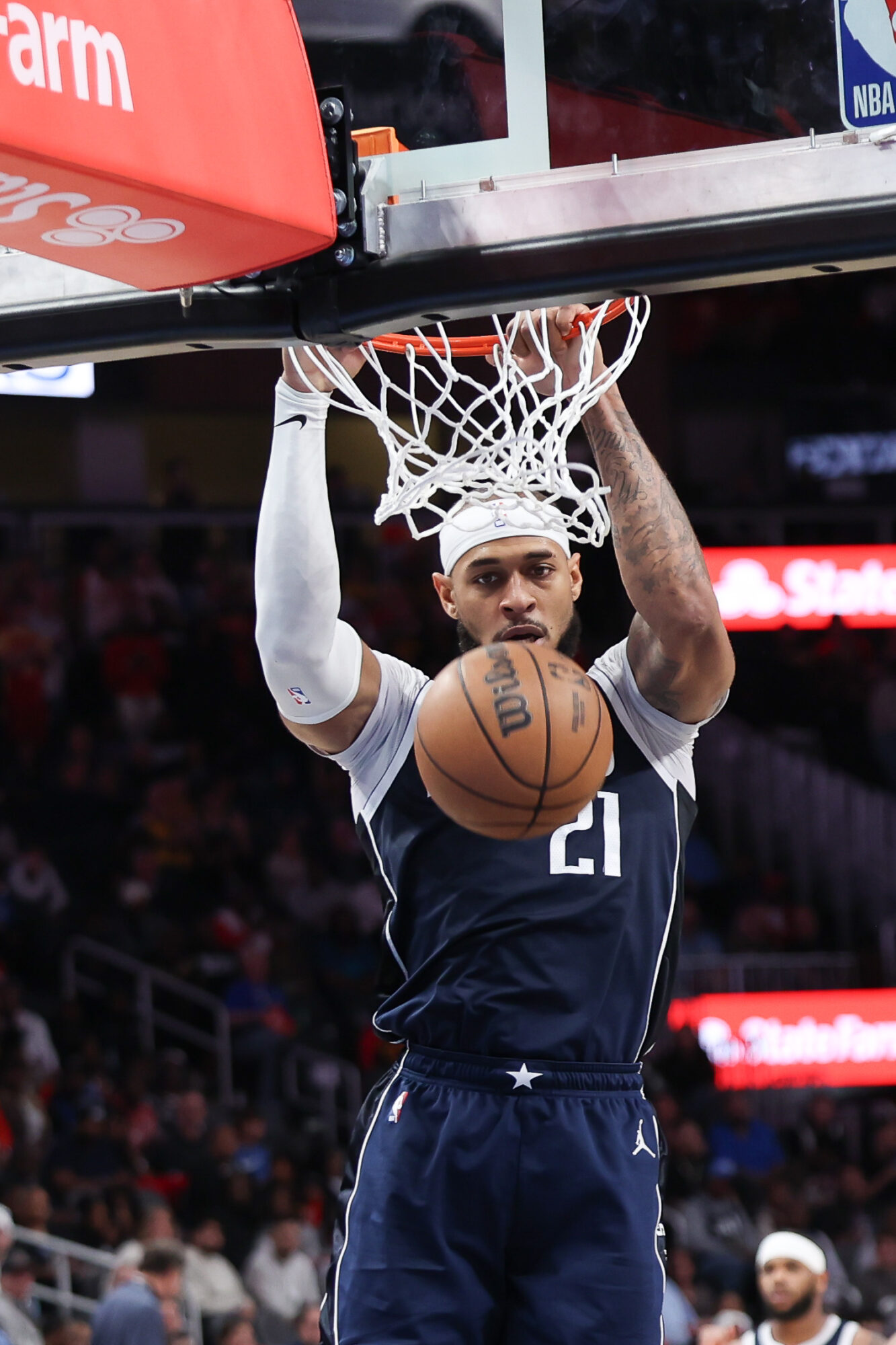 Basketball player in a dark uniform dunks the ball, with a basketball hoop and net above him.