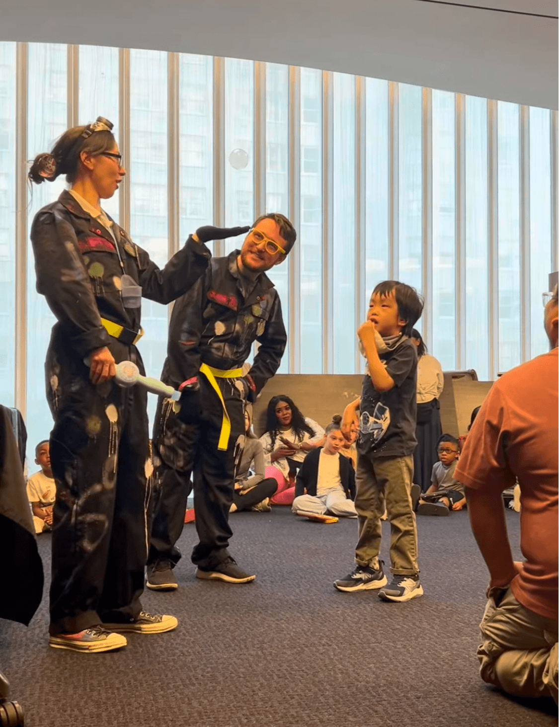Group of children and adults in a room with large windows, some children sitting on the floor, one child talking to adults.
