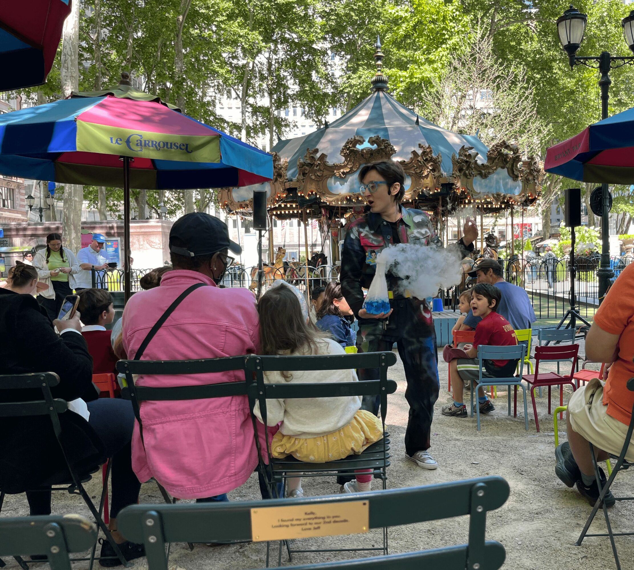 People sitting around a carousel in a park, with colorful umbrellas and trees in the background.