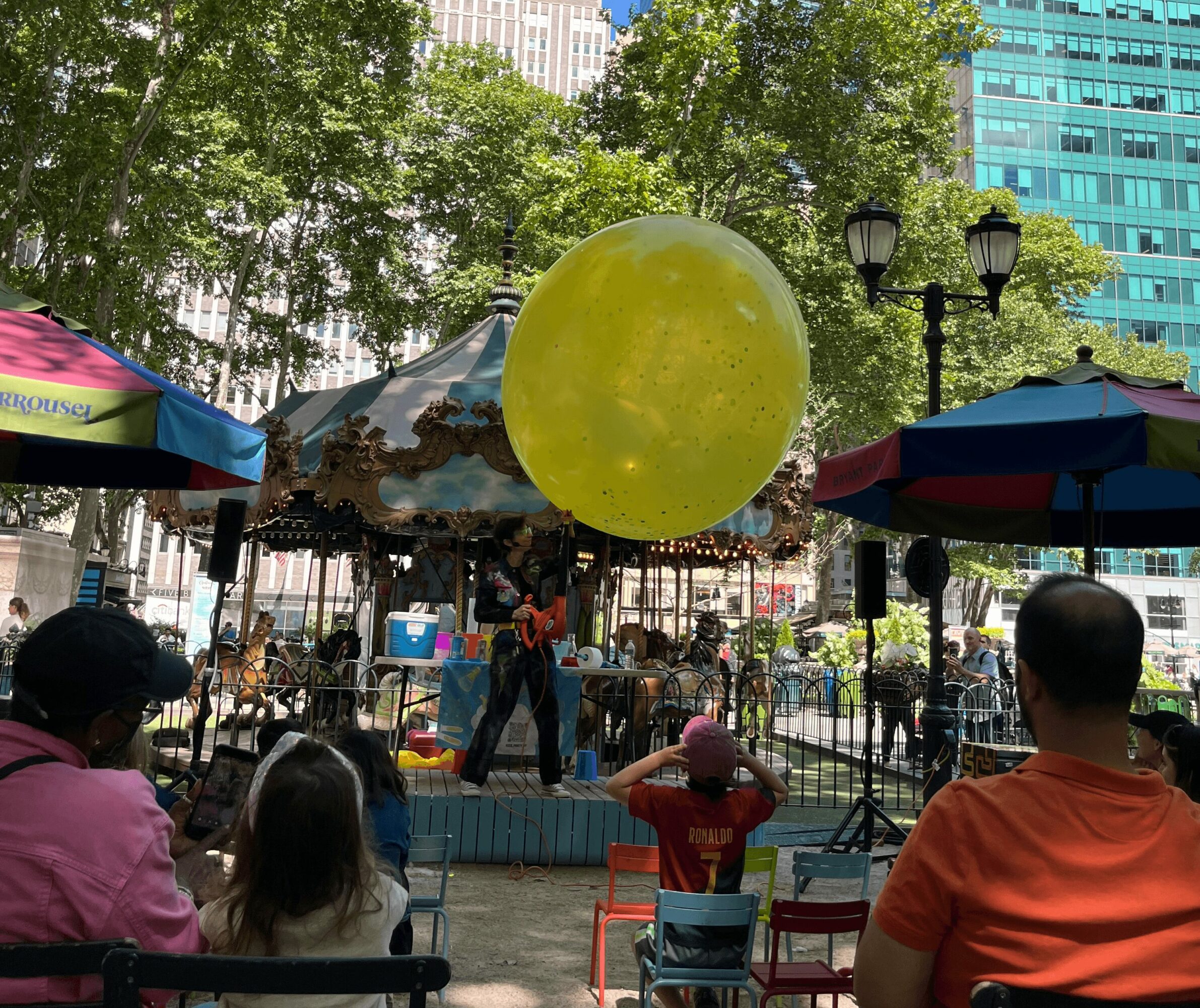 People watching a performer with a large yellow balloon in a park with trees, umbrellas, and buildings in the background.