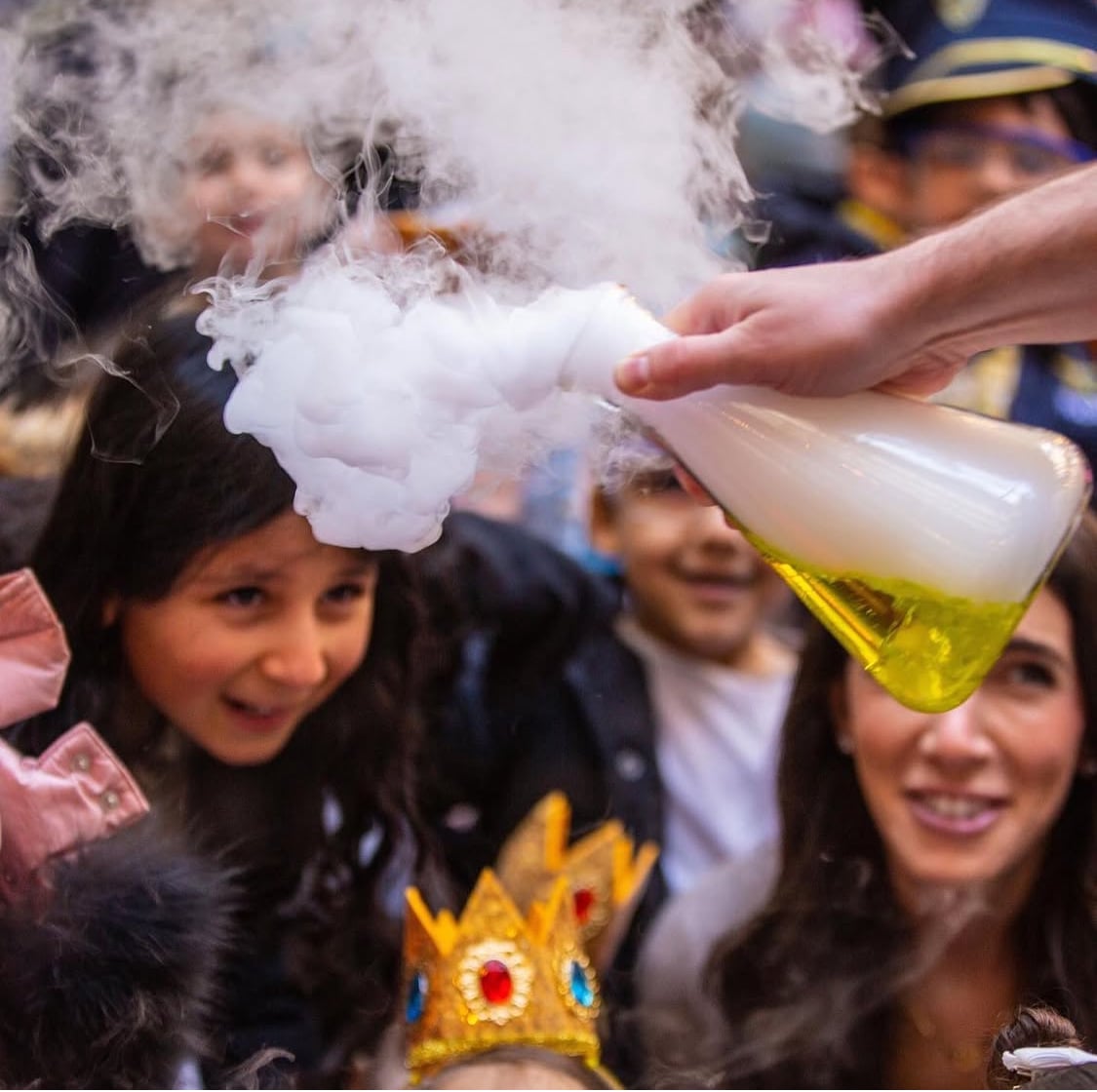 Person holding a flask with yellow liquid, releasing white smoke, surrounded by smiling children and a woman, during a science demonstration.