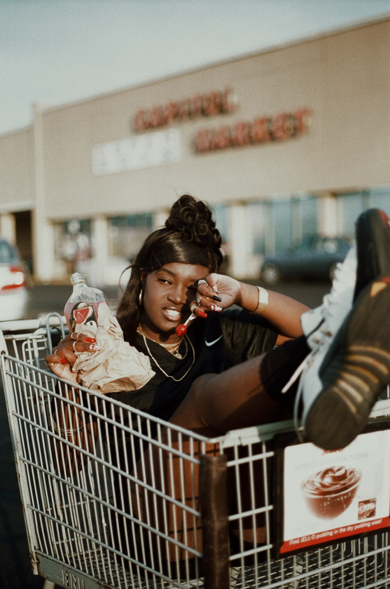 Person with dark hair in a bun sitting in a shopping cart outside store, holding a phone, with store and cars in background.