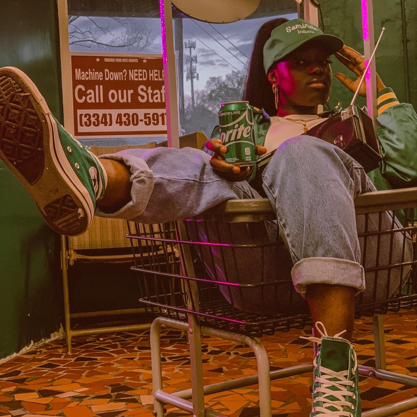 Person sitting in a shopping cart with legs extended, holding a Sprite can, wearing sneakers and a cap, inside a store.