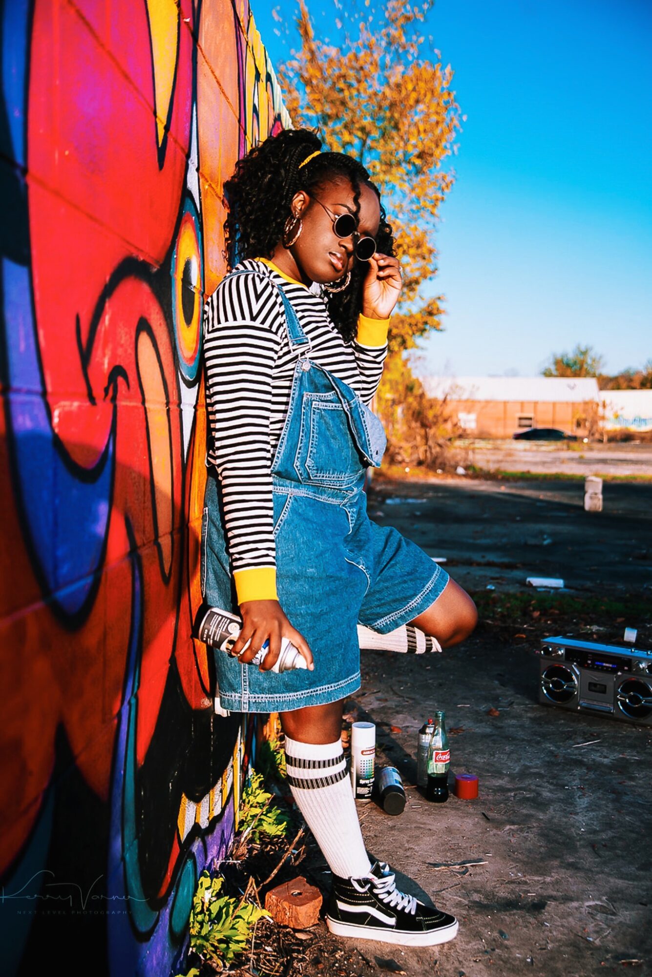 Young woman with curly hair and sunglasses leaning against a colorful graffiti wall outdoors, holding a phone and a bag.