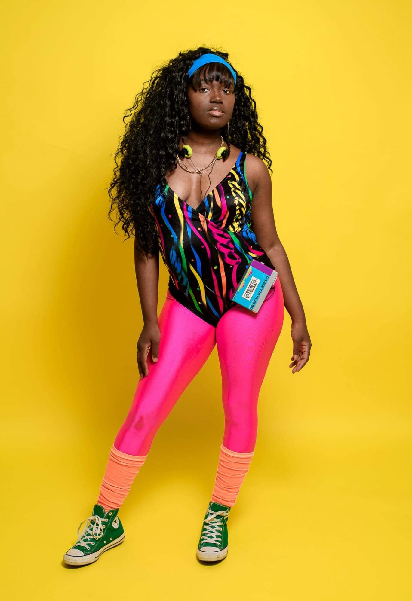Young girl with long curly hair wearing colorful outfit and accessories, standing against yellow background.