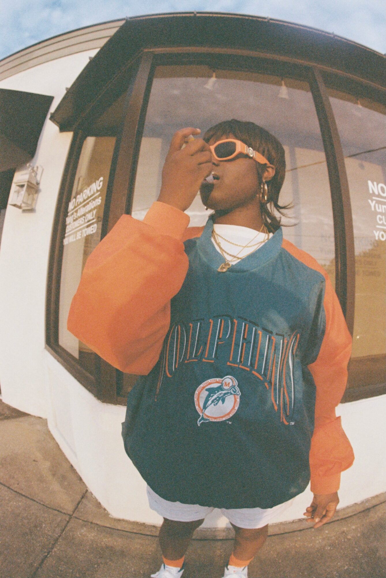 Person wearing sunglasses and a Miami Dolphins jacket, standing outside a building with large windows, looking to the side.