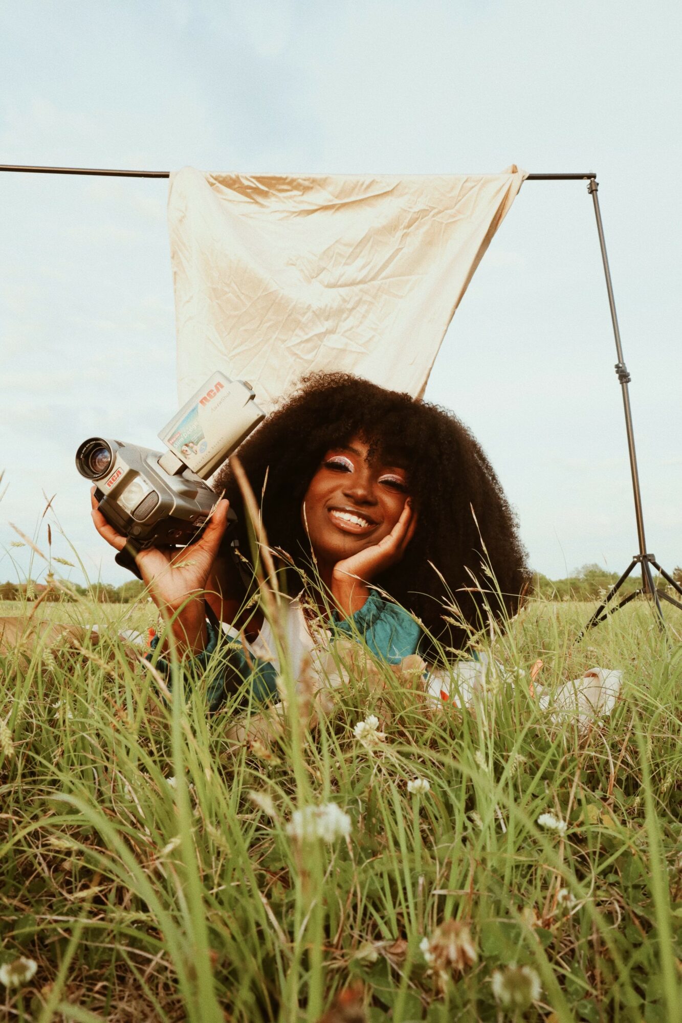 Woman with curly hair lying in grass, holding a video camera, smiling, outdoors with a backdrop and grass.