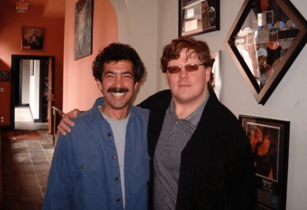 Two men stand close together, smiling, in a room with framed pictures on the wall.