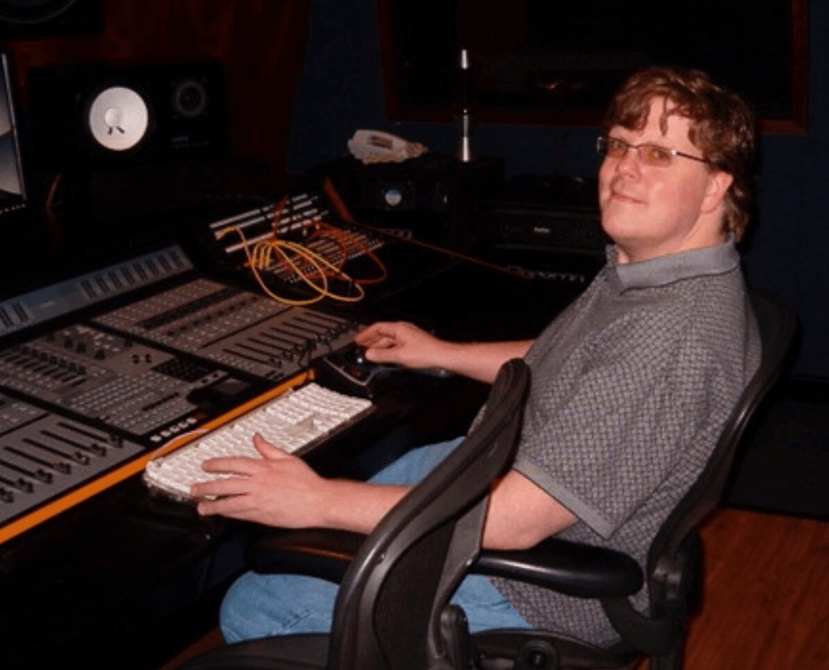 Person with glasses sitting at a mixing console in a recording studio, smiling, with audio equipment around.