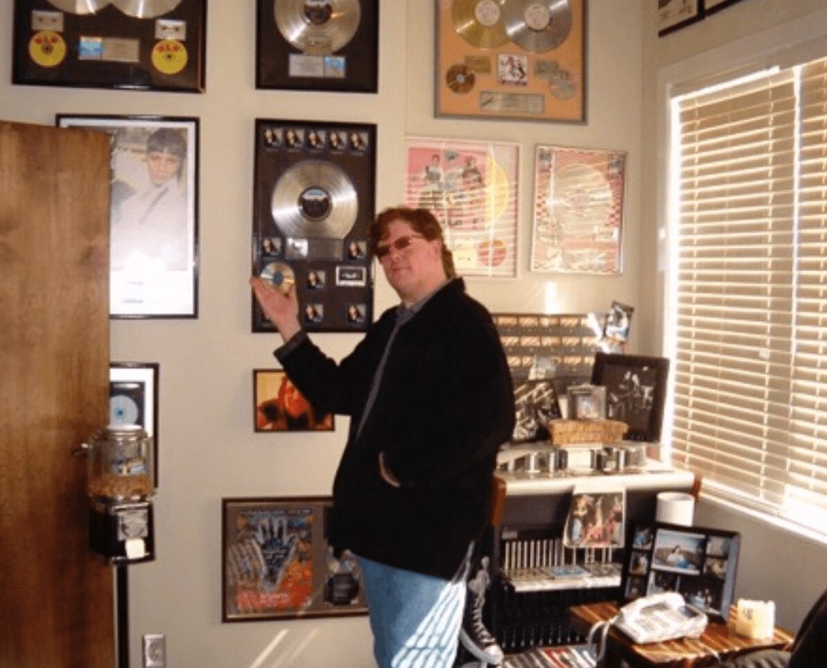Person standing in a room with music awards and framed pictures on the wall, holding a small object, near a window with blinds.