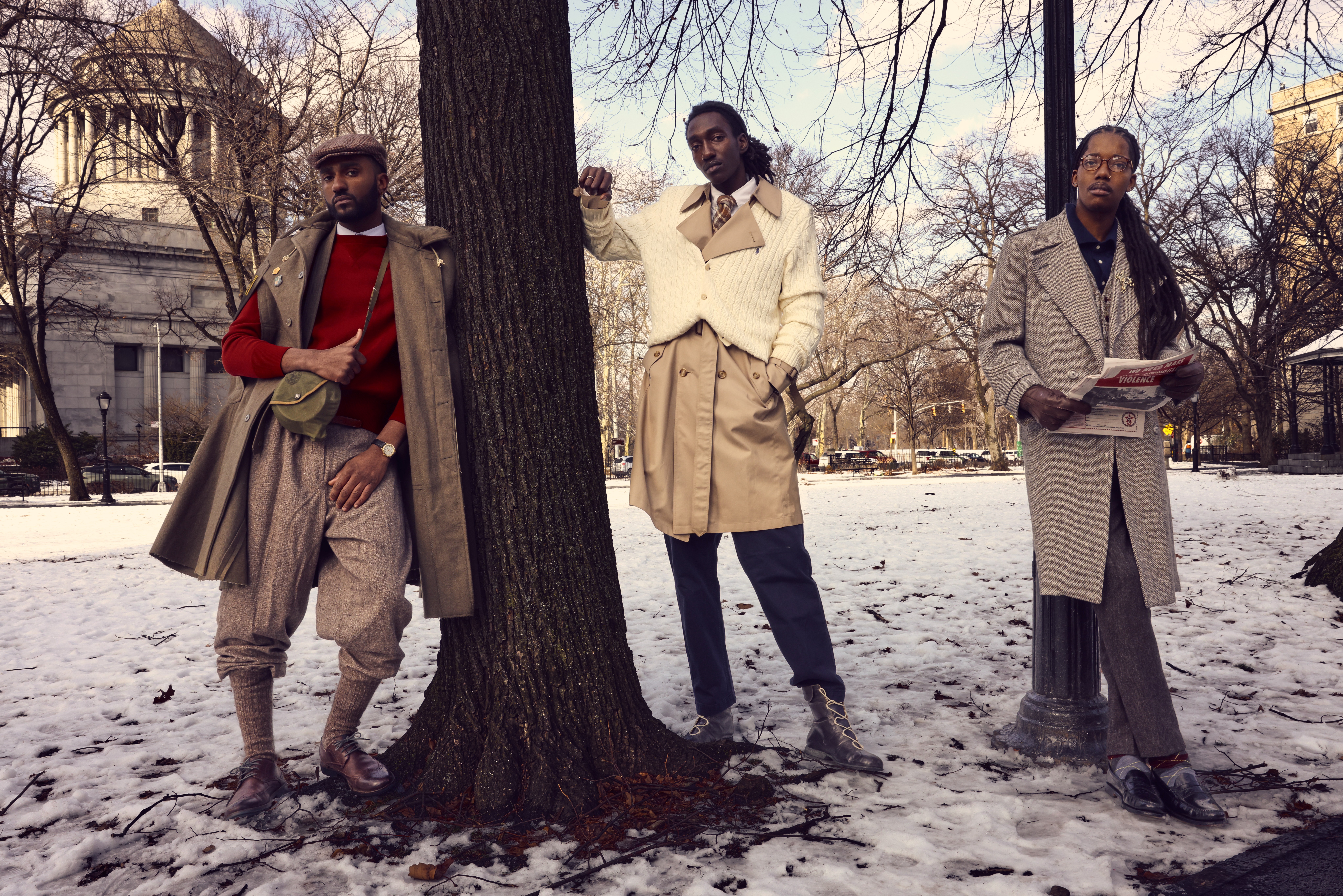 Three people standing outdoors in a snowy park with trees and buildings in the background.