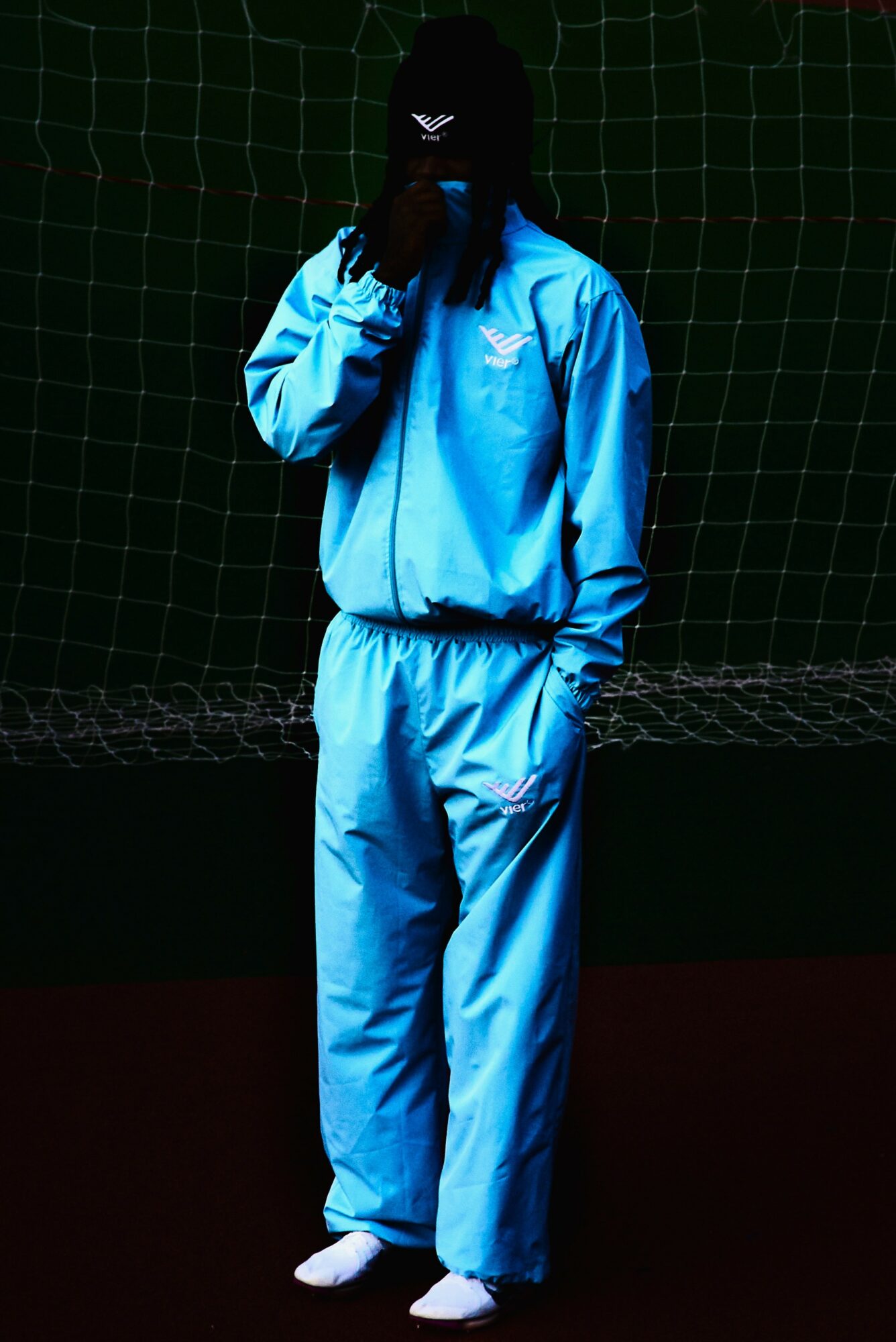 Person in blue tracksuit standing in front of a net, face covered, holding a phone, dark background.