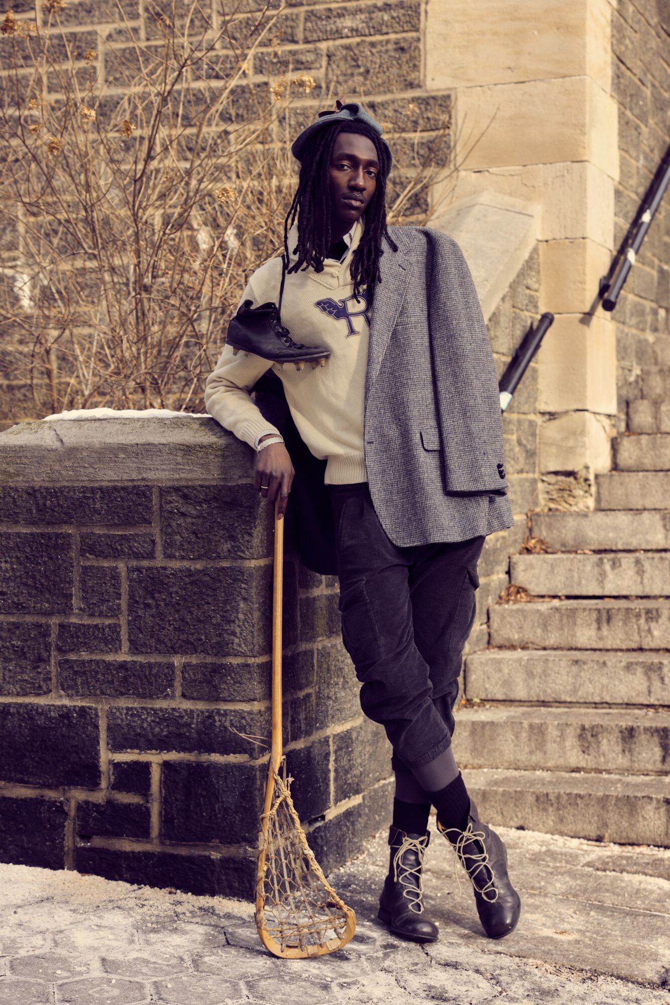 Person with dreadlocks leaning on a stone wall holding a lacrosse stick, wearing a gray coat and black boots.