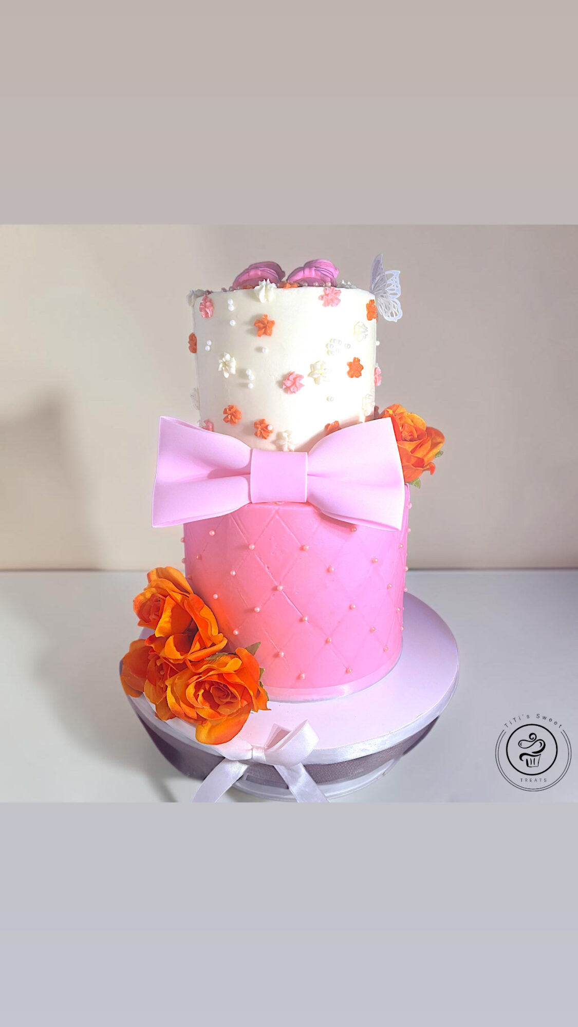 Two-tiered cake with pink and white fondant, decorated with flowers and a large pink bow, on a white cake board.