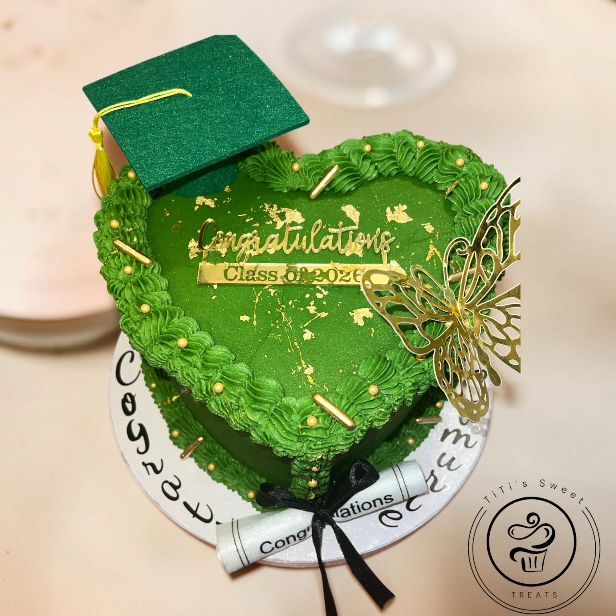 Heart-shaped green graduation cake with a cap, butterfly decoration, and a scroll reading 'Congratulations'.