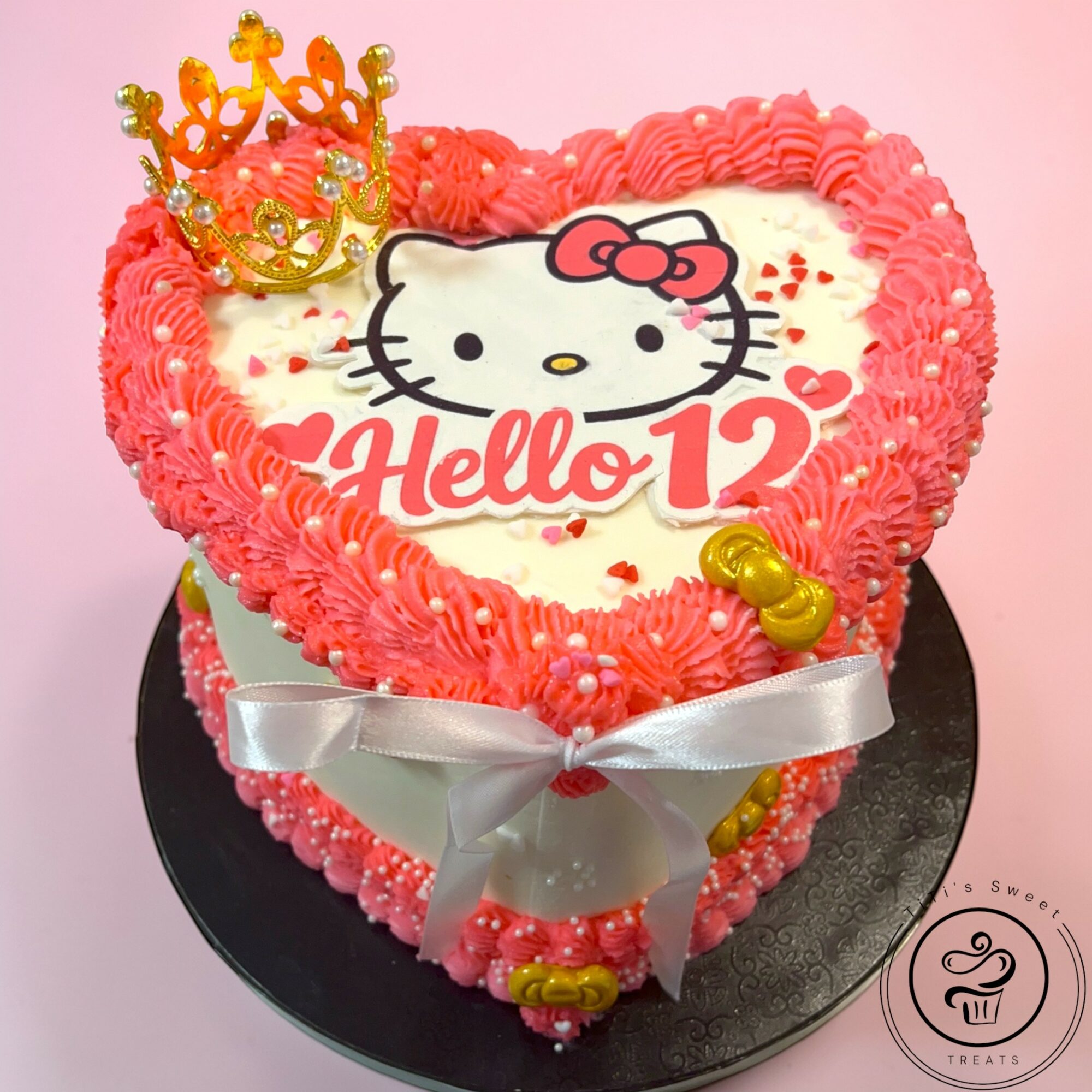 Heart-shaped cake with Hello Kitty design, pink and white frosting, a gold crown, and a silver ribbon bow, on a black base.
