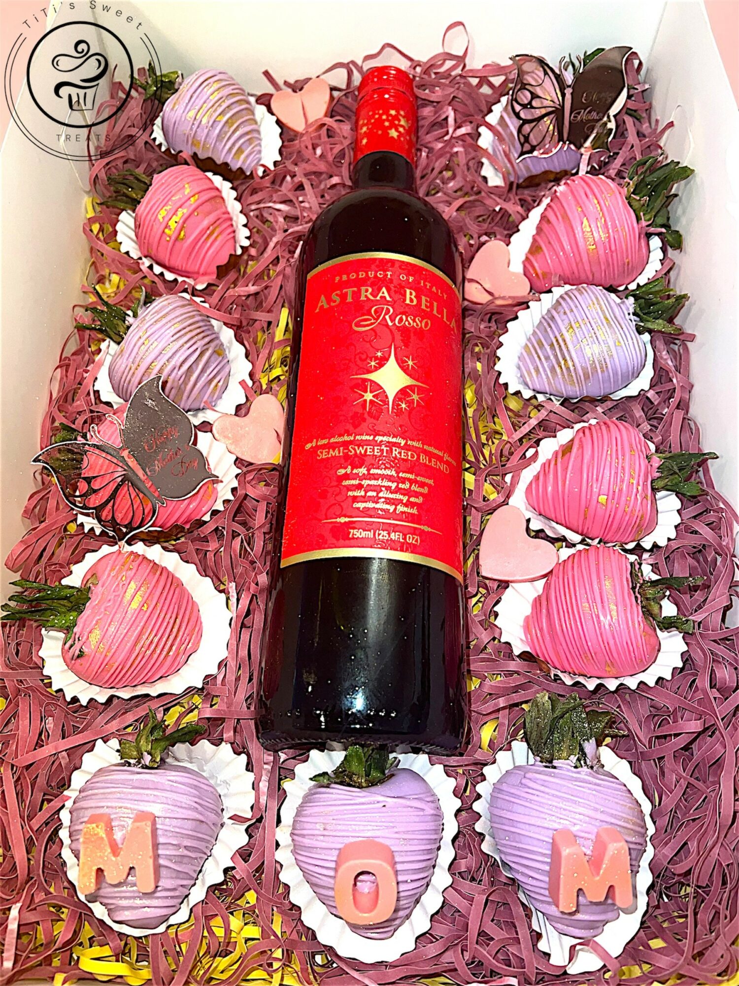 A bottle of red wine surrounded by pink and purple heart-shaped chocolates with the word 'MOM' on three of them, pink shredded paper.