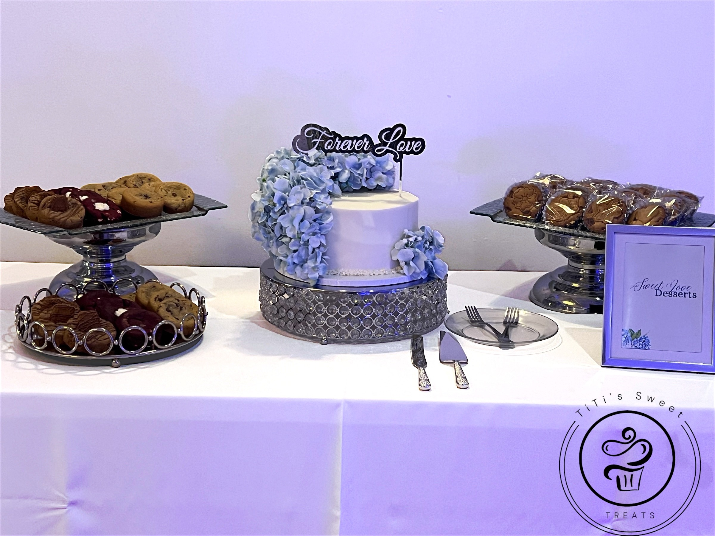 Cake with blue flowers and a topper, surrounded by cookies and chocolates on a table, with a sign and utensils.