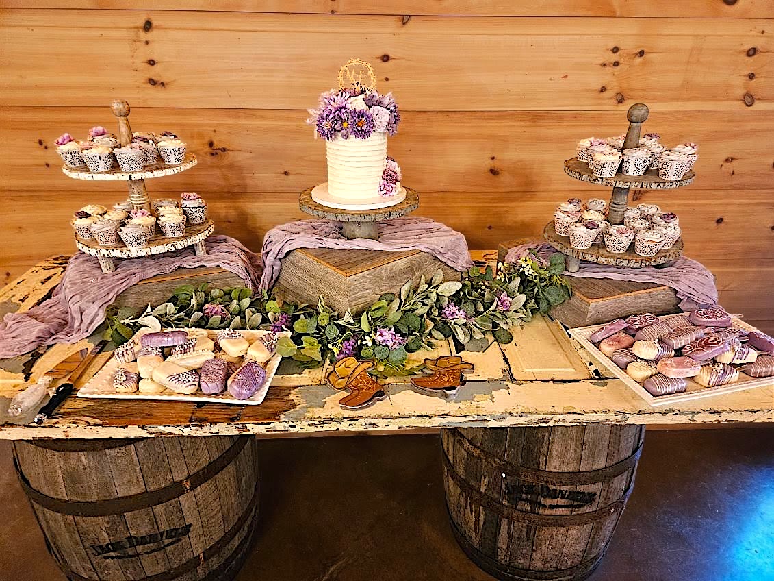 Table with cupcakes, a cake with flowers, and decorative items against a wooden wall, with a rustic wooden table and barrels.