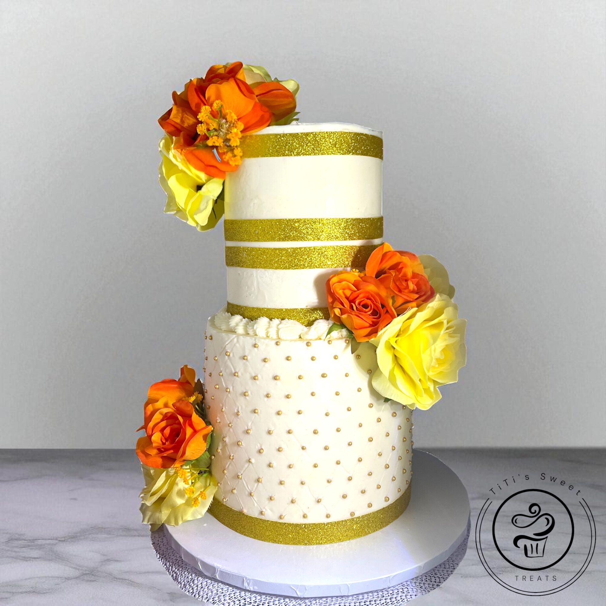 Two-tiered cake decorated with orange and yellow flowers, gold bands, and a white base, on a marble surface.
