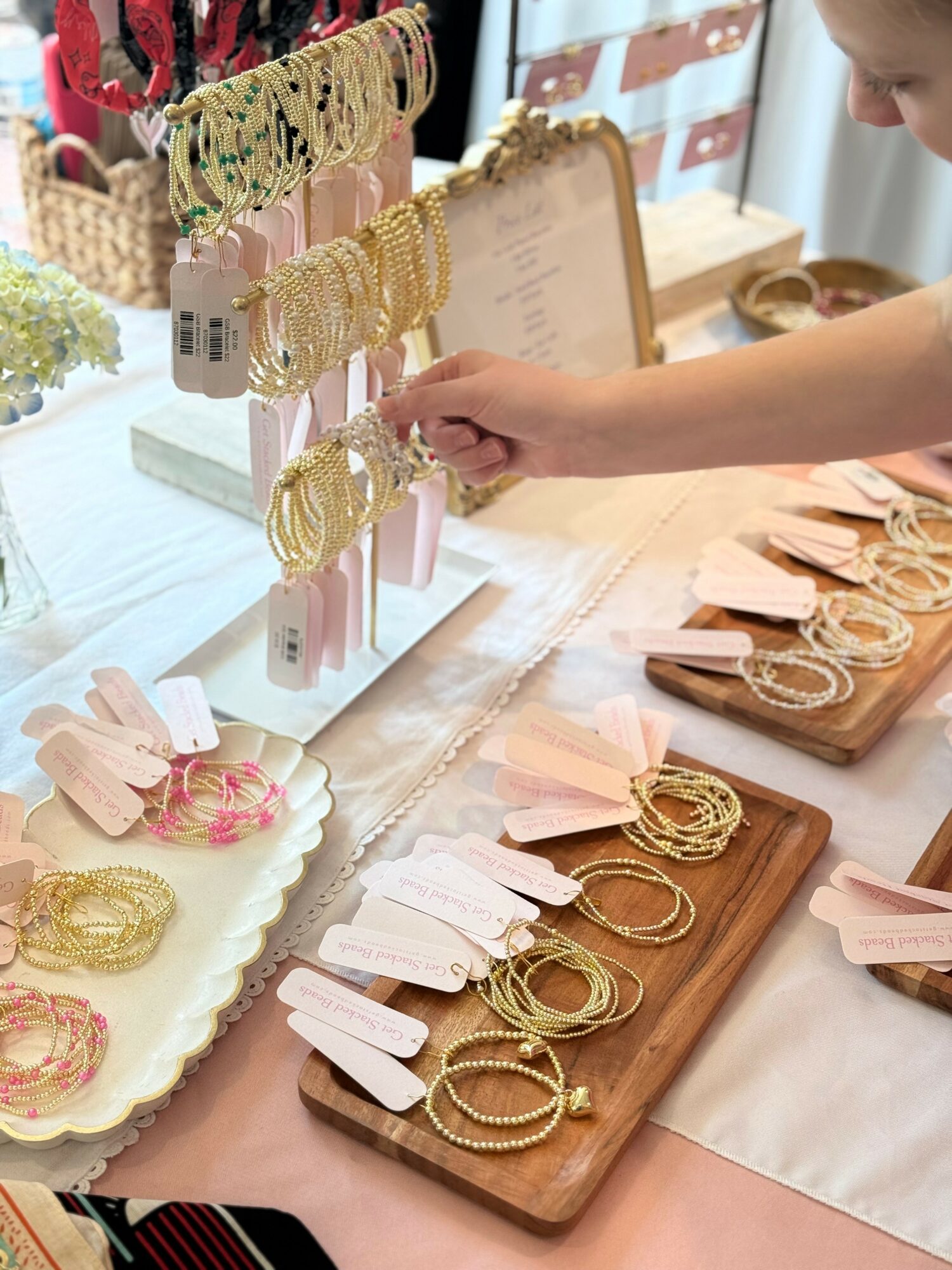 Person browsing jewelry display with necklaces and bracelets on wooden and ceramic trays, in a well-lit shop.