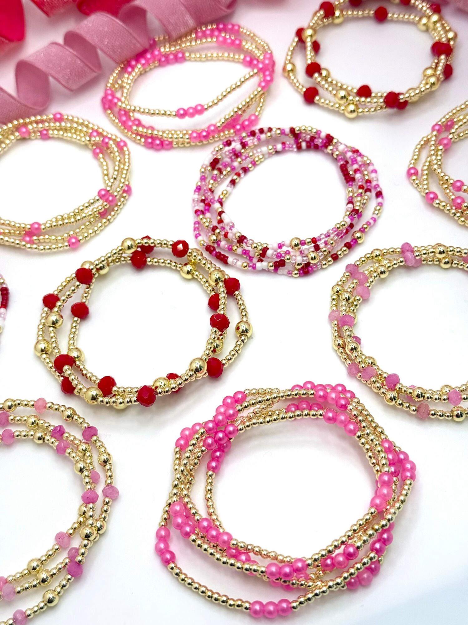 Multiple beaded bracelets in pink, red, and white colors arranged on a white surface with pink ribbon in background.
