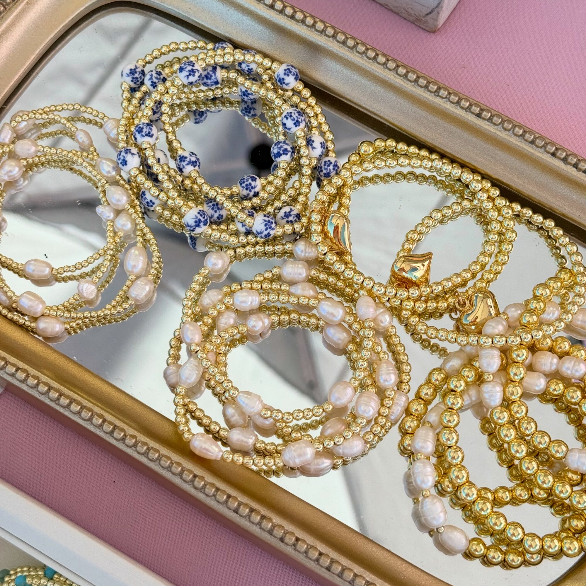 Multiple pearl and gold-colored jewelry pieces, including bracelets and necklaces, arranged on a mirrored tray with a pink background.