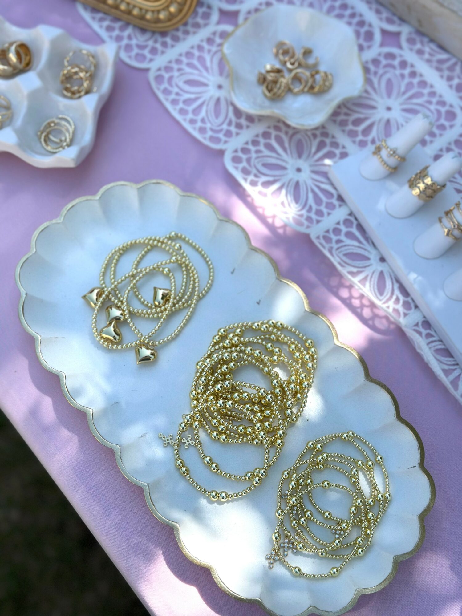 Gold jewelry including necklaces and bracelets displayed on white dishes and a pink tablecloth, with lace and decorative items nearby.