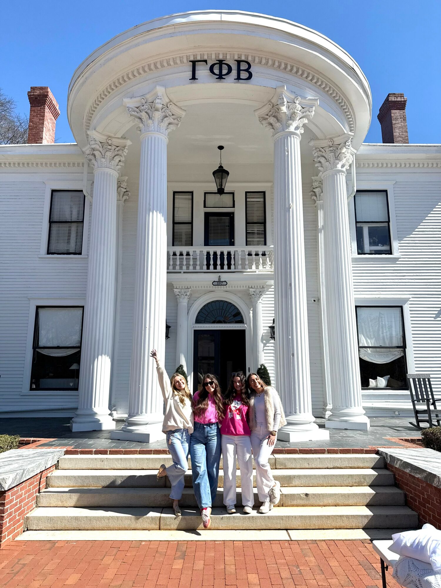 Four people stand on steps in front of a large white building with columns and Greek letters, under a clear blue sky.