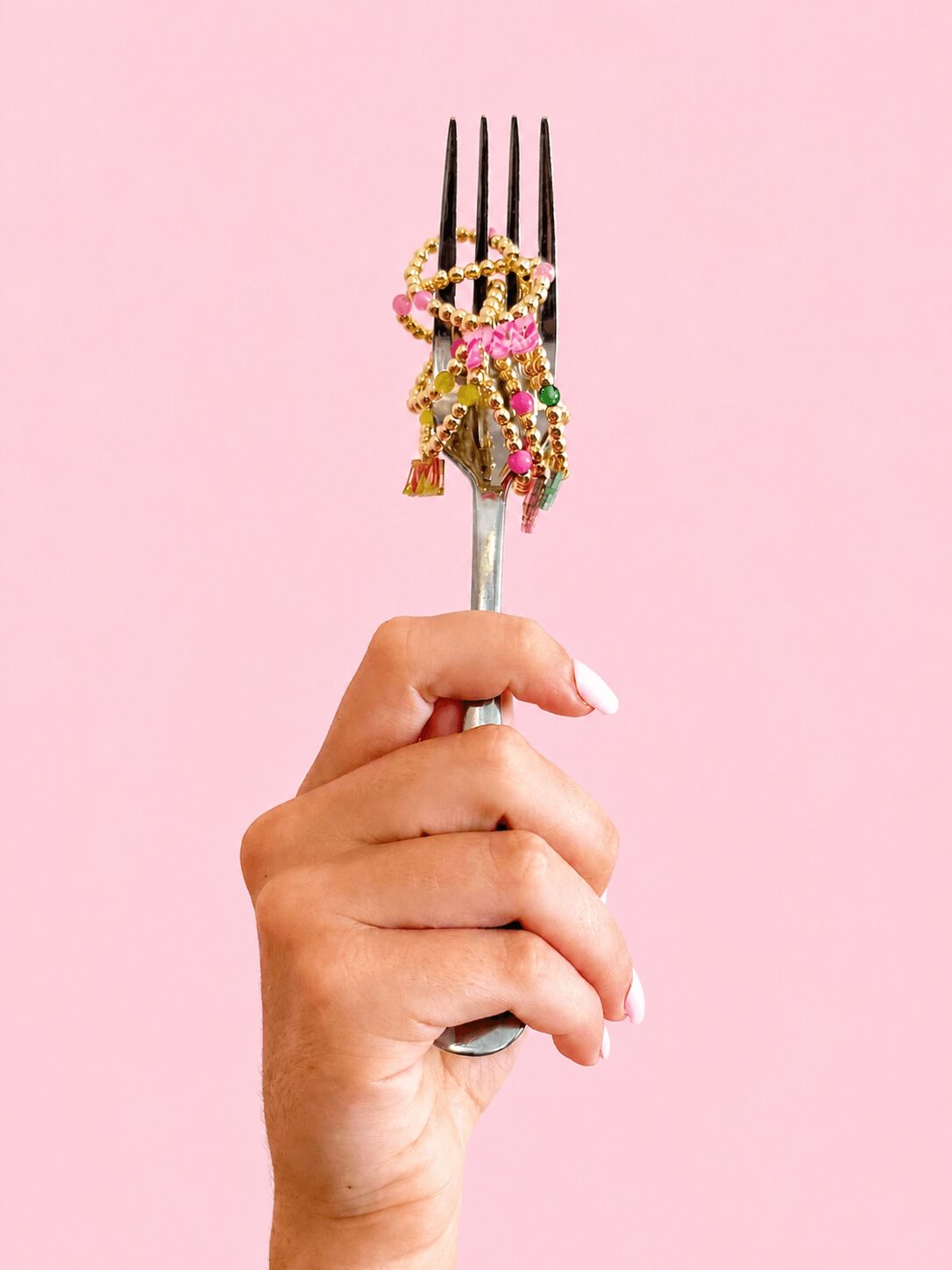 Hand holding a fork with colorful jewelry against a pink background.